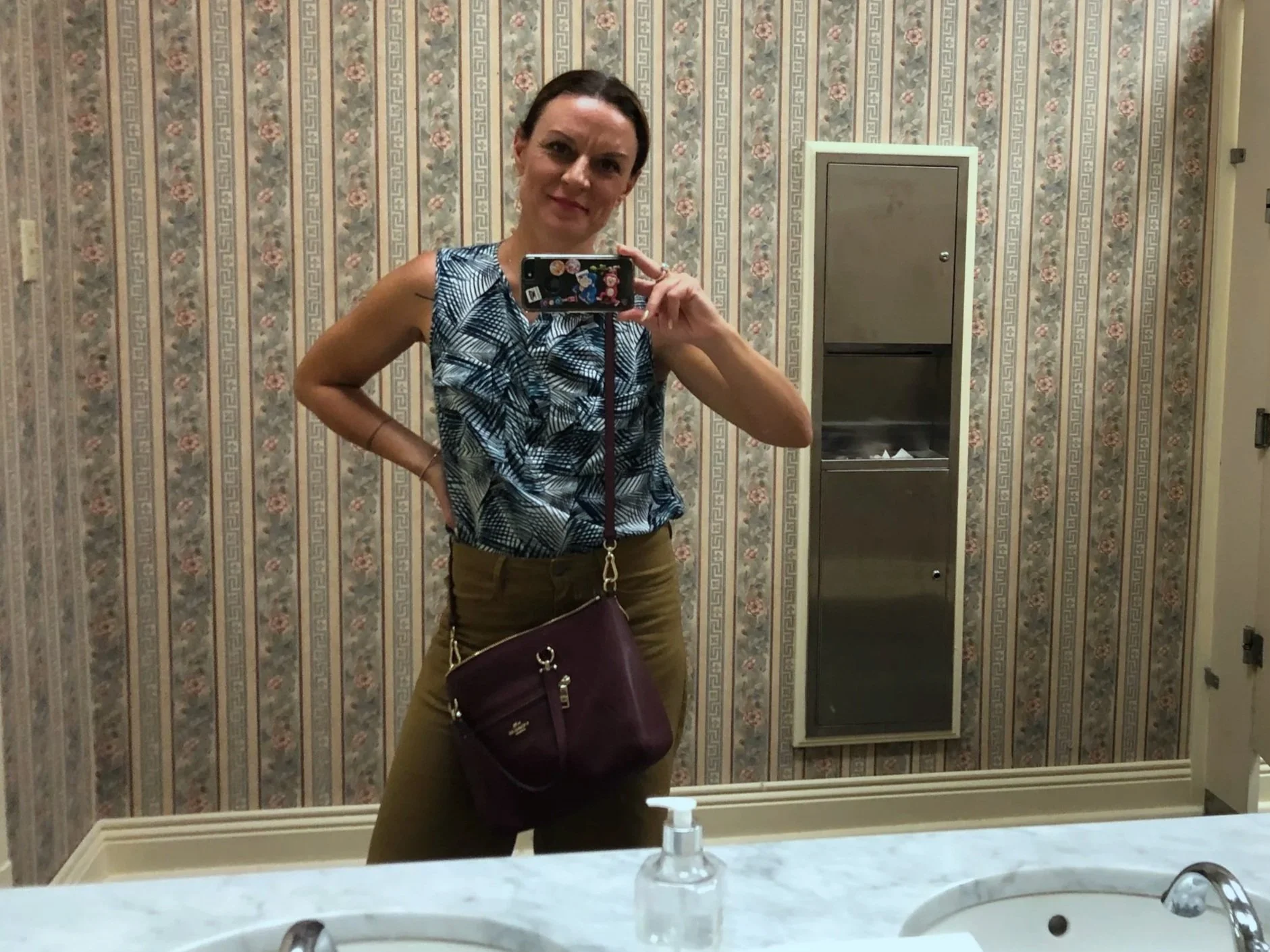 A woman taking a mirror selfie in a bathroom, wearing a sleeveless patterned top and khaki pants, with a purple purse hanging from her shoulder. The bathroom has floral wallpaper, a marble countertop with a sink, and a soap dispenser. There is a gray cabinet on the wall behind her.