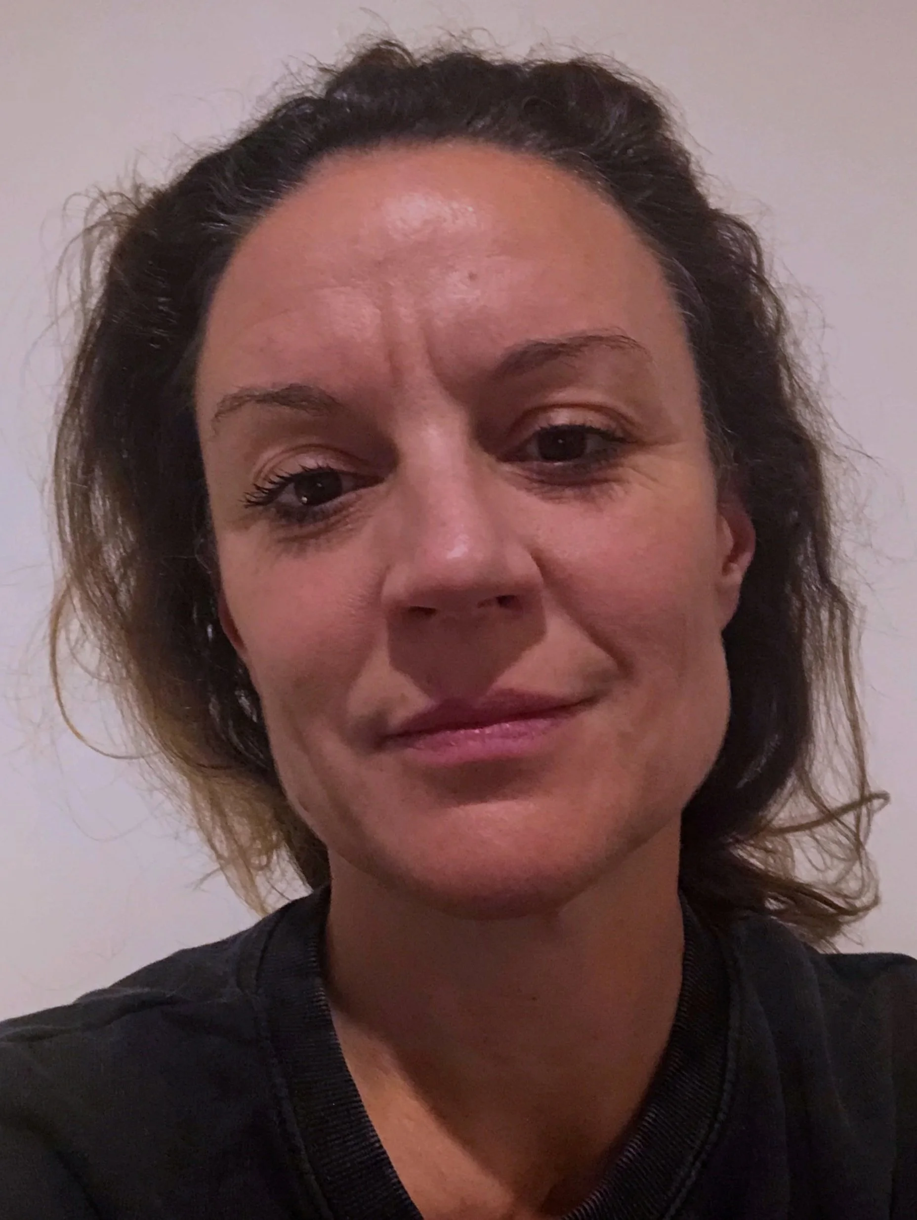 Close-up of a woman with curly hair and light makeup, wearing a black top, against a plain wall background.