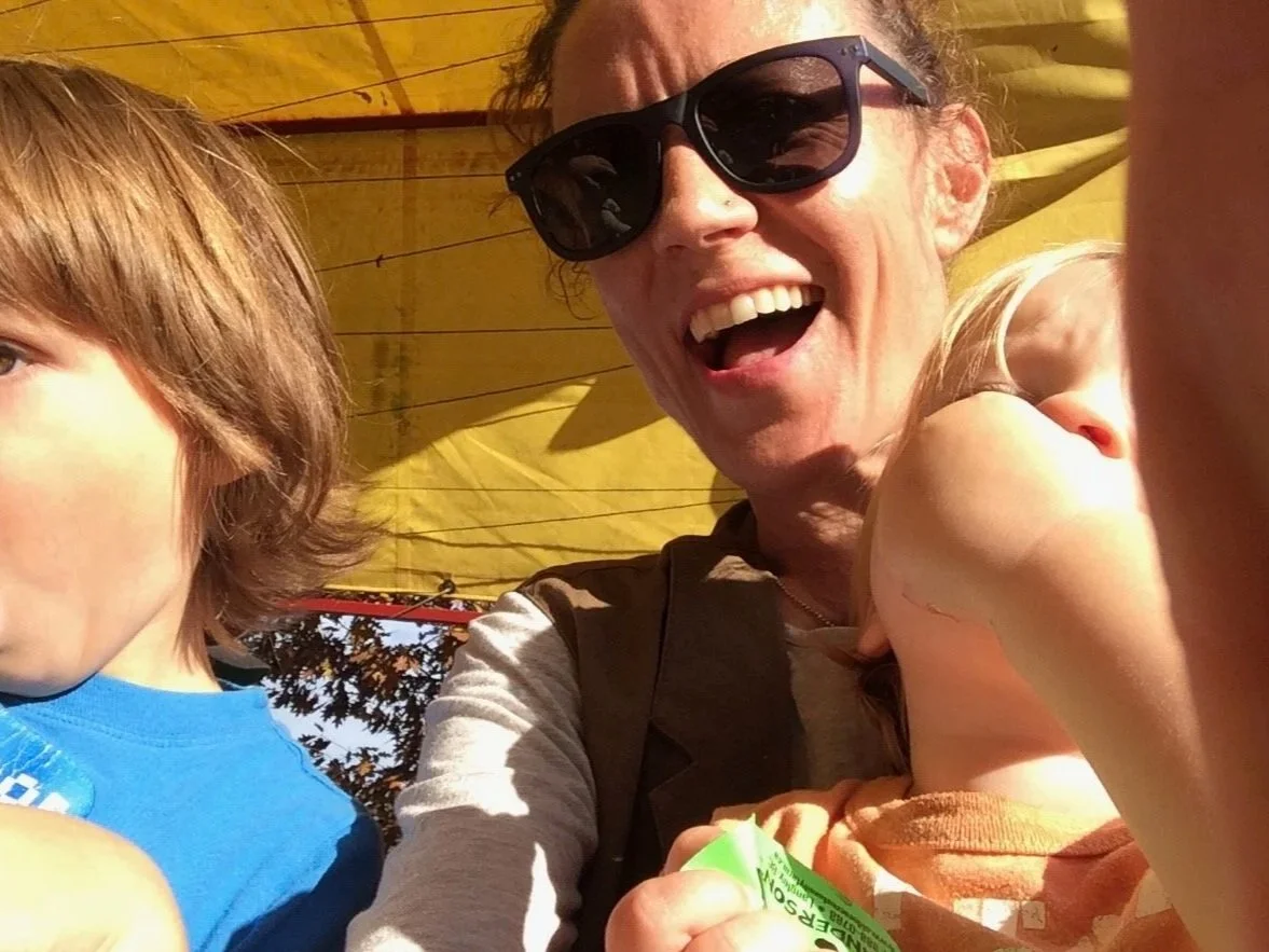 A woman wearing sunglasses smiles with two young children: one with brown hair in a blue shirt and one with blonde hair in an orange shirt, outdoors under a yellow canopy with trees in the background.