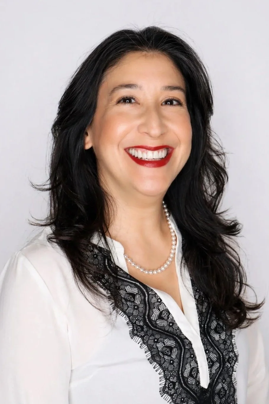 A smiling woman with dark wavy hair, wearing a white blouse with black lace detail and a pearl necklace, standing against a light background.