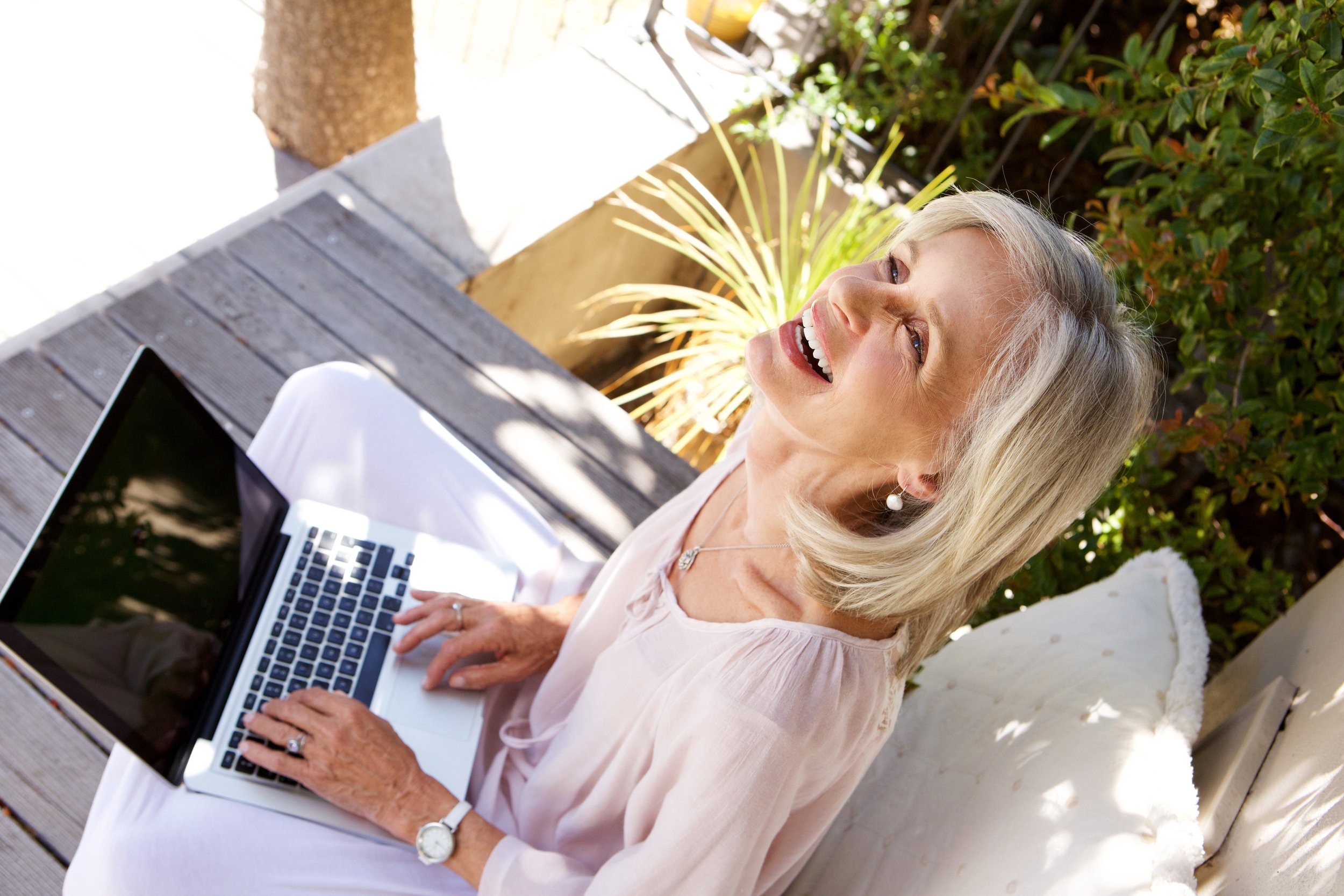 Older woman sitting outdoors on a patio, working on a laptop, smiling, surrounded by plants and sunlight.