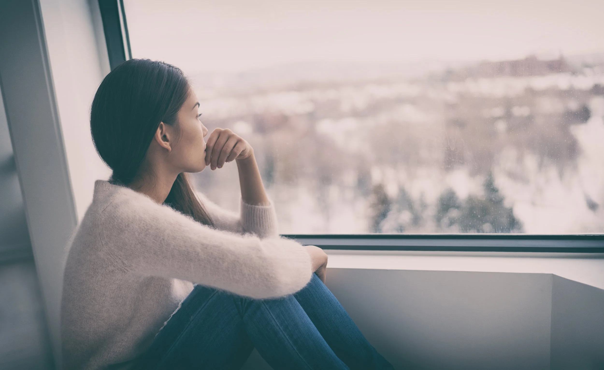 A woman sitting by a window, looking outside thoughtfully on a cloudy day.