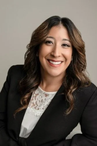 A woman with long brown hair smiling, wearing a black blazer and a white lace top, against a plain gray background.