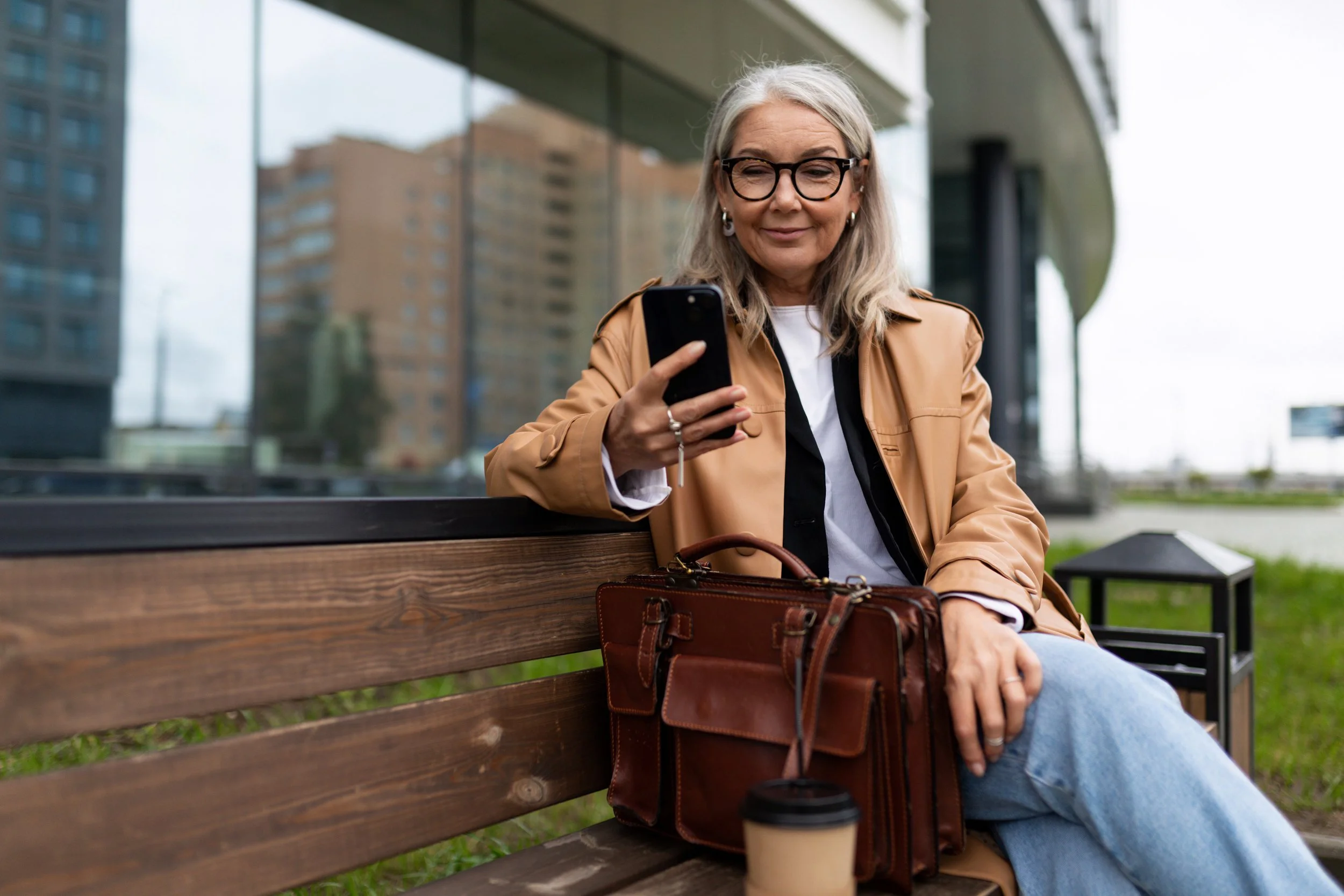 Older woman with glasses and gray hair sitting on a park bench, looking at her phone, with a leather bag and a paper coffee cup beside her, in an urban setting with modern buildings in the background.