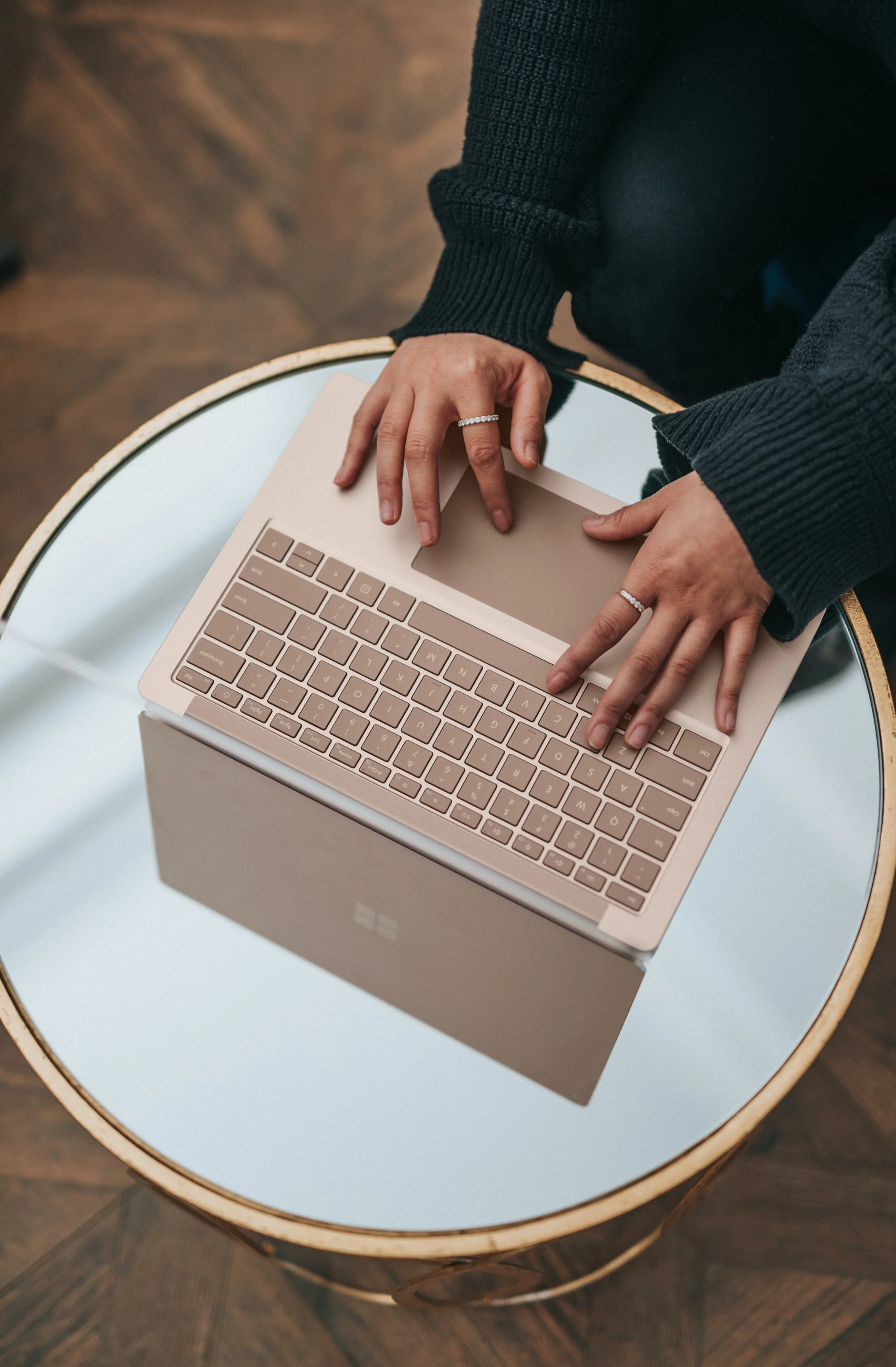 A person using a beige laptop with a reflective screen, sitting at a round glass-topped table with a gold trim.