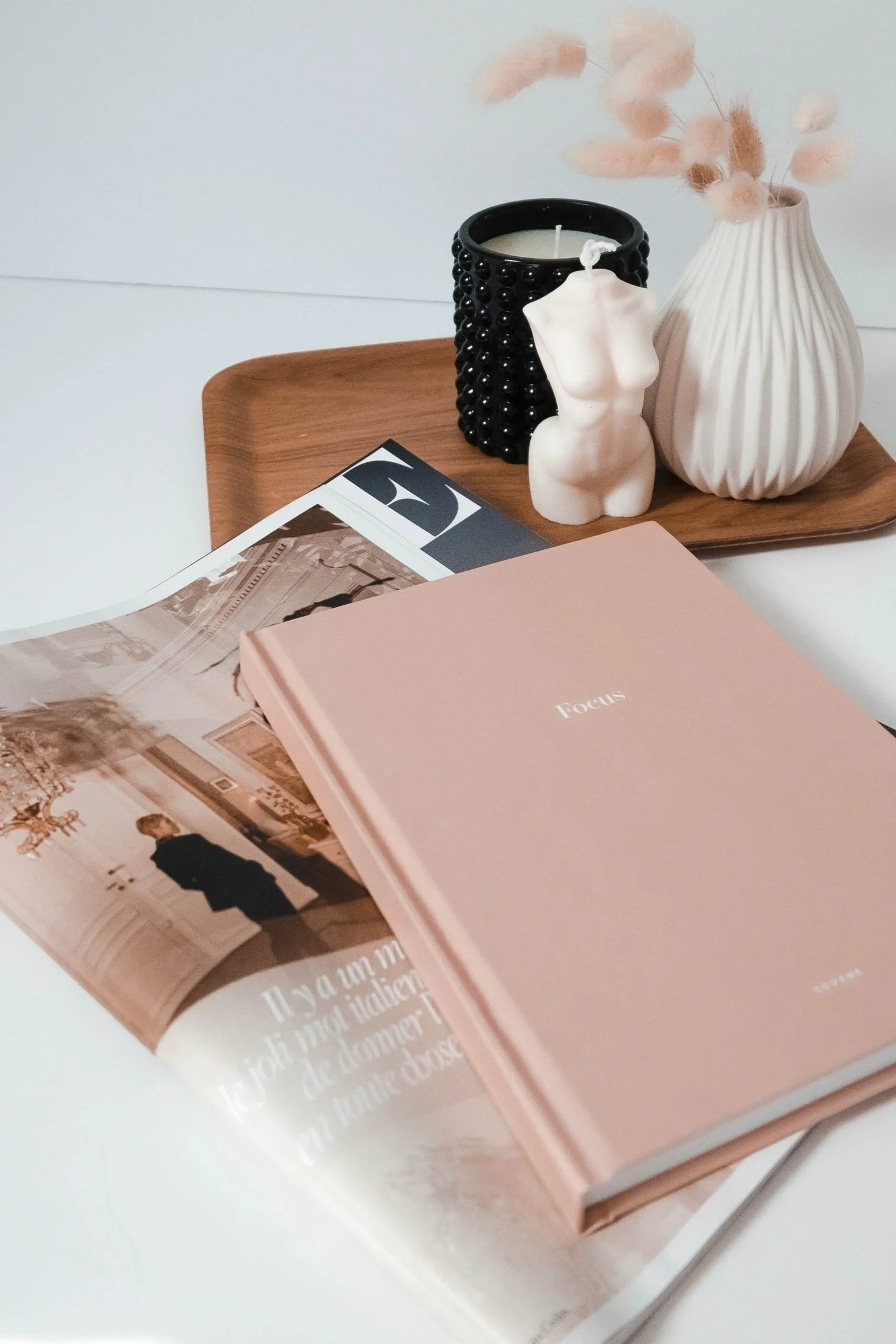 Flat lay of a wooden tray with decorative objects including a black textured candle, a small white torso sculpture, a white pleated vase with dried pink pampas grass, a pastel pink book titled "Focus," and an open magazine with a photo of a person in a black coat, on a white surface.