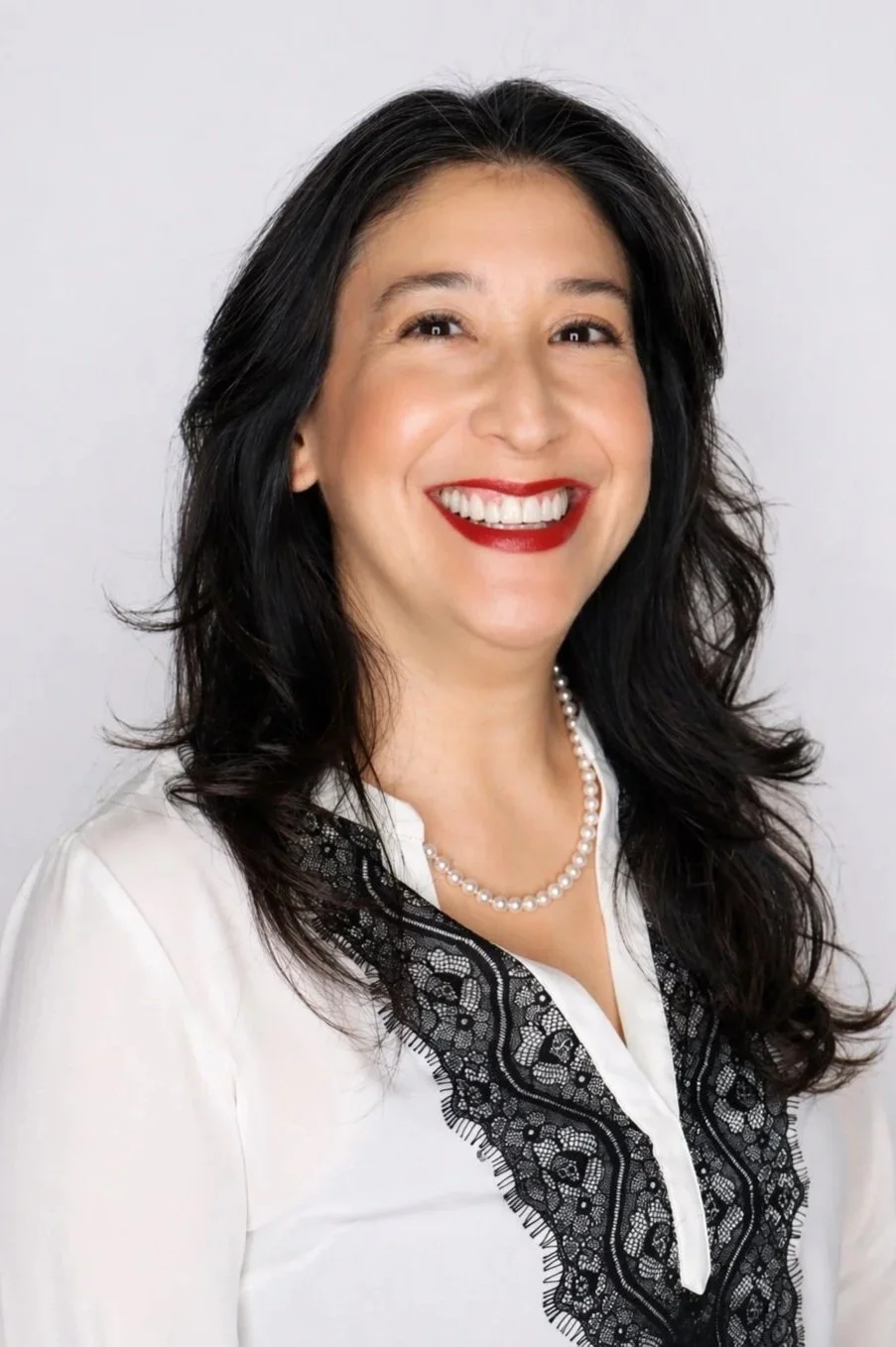 A woman with long black hair, wearing a white blouse with black lace details and a pearl necklace, smiling against a plain white background.