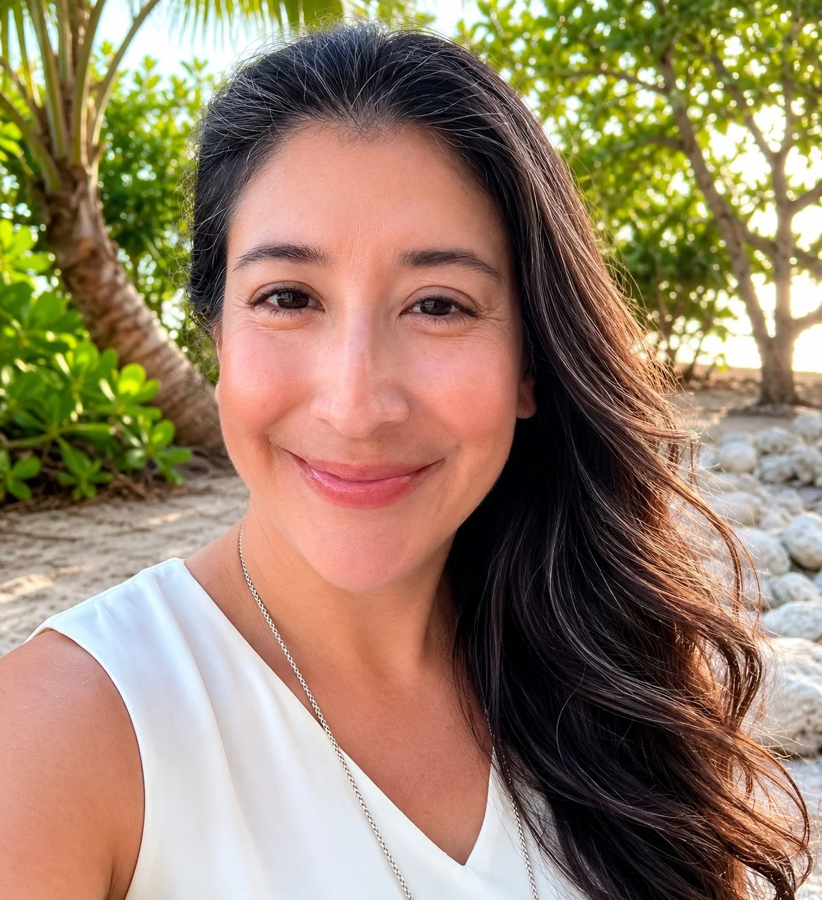 A woman smiling outdoors with tropical plants and trees in the background during sunset.