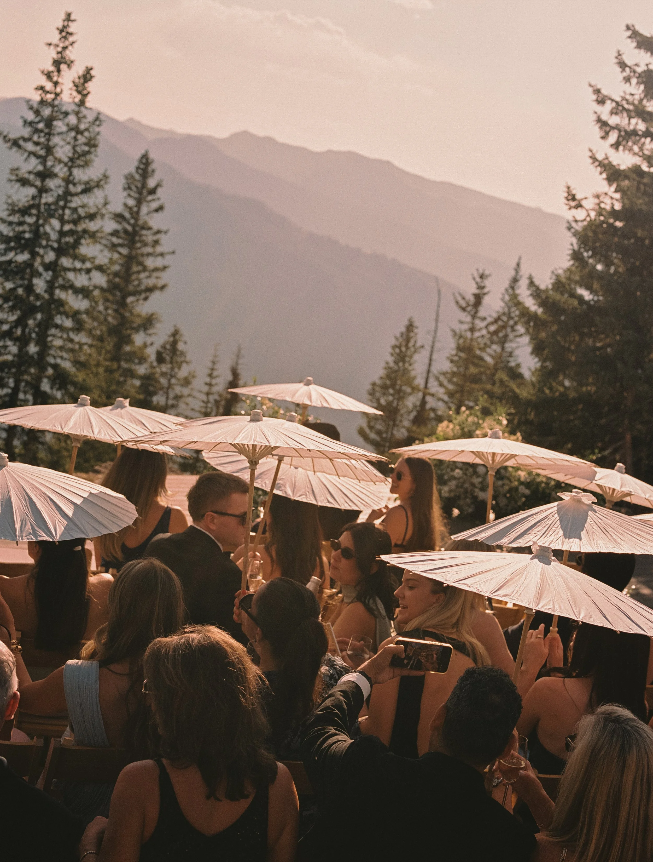 Crowd of people at outdoor event during sunset, holding pink umbrellas, with trees and mountains in background.