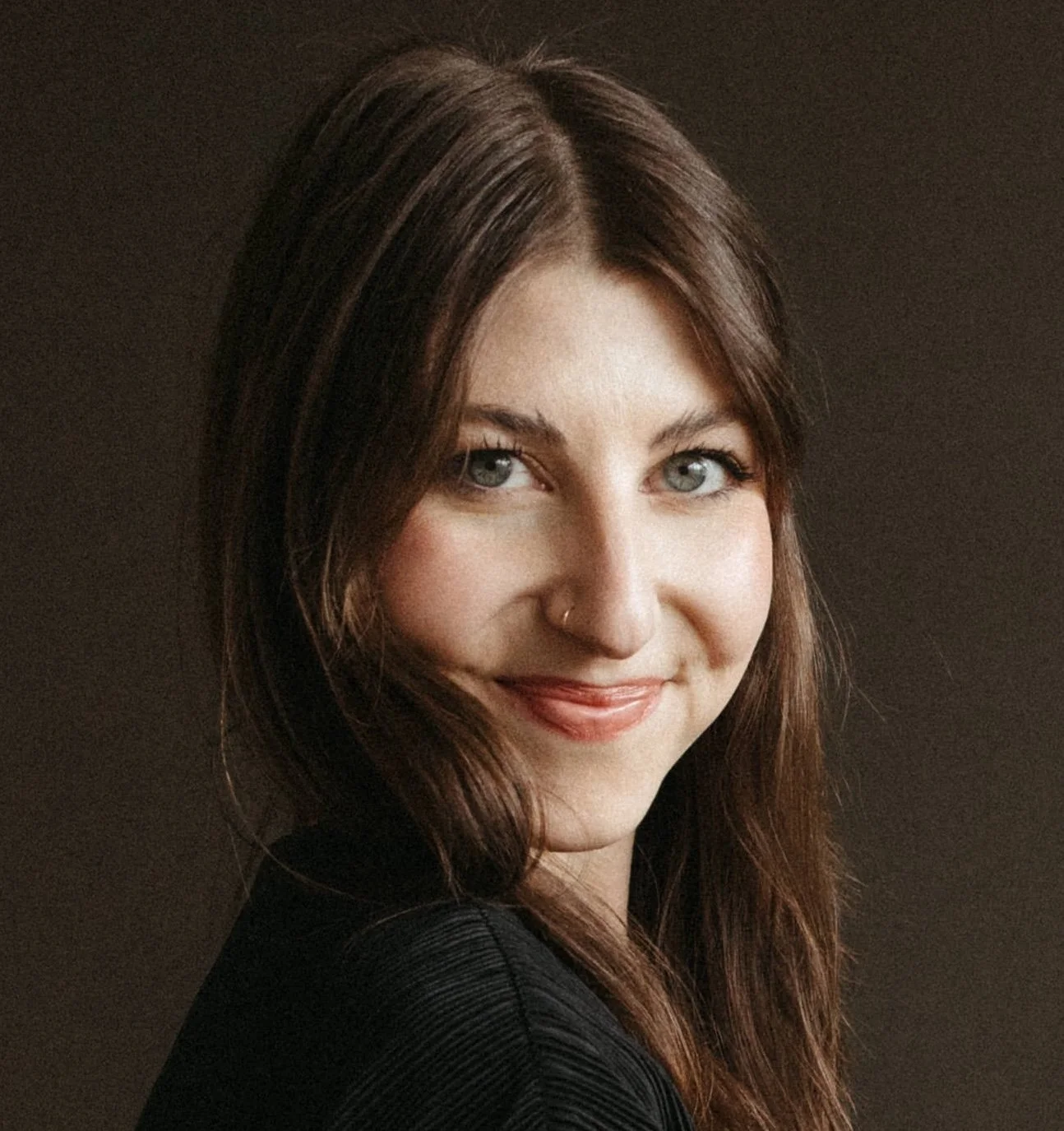 Portrait of a woman with long brown hair and blue eyes, smiling against a plain dark background.