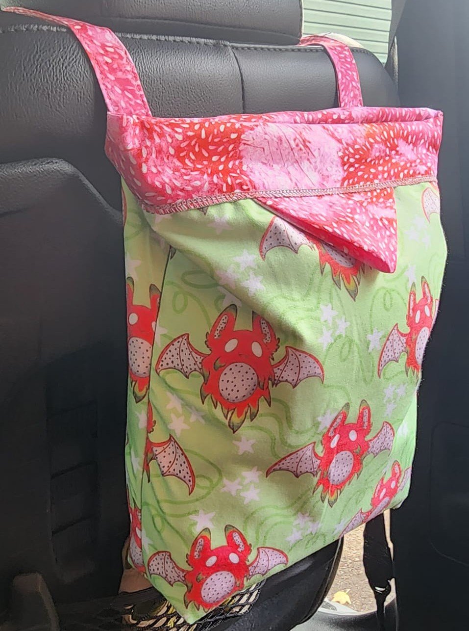 A colorful fabric bag with a pattern of red cartoon dragons with bat-like wings on a green background, hanging on a car seat. Car Snap closed trash can.