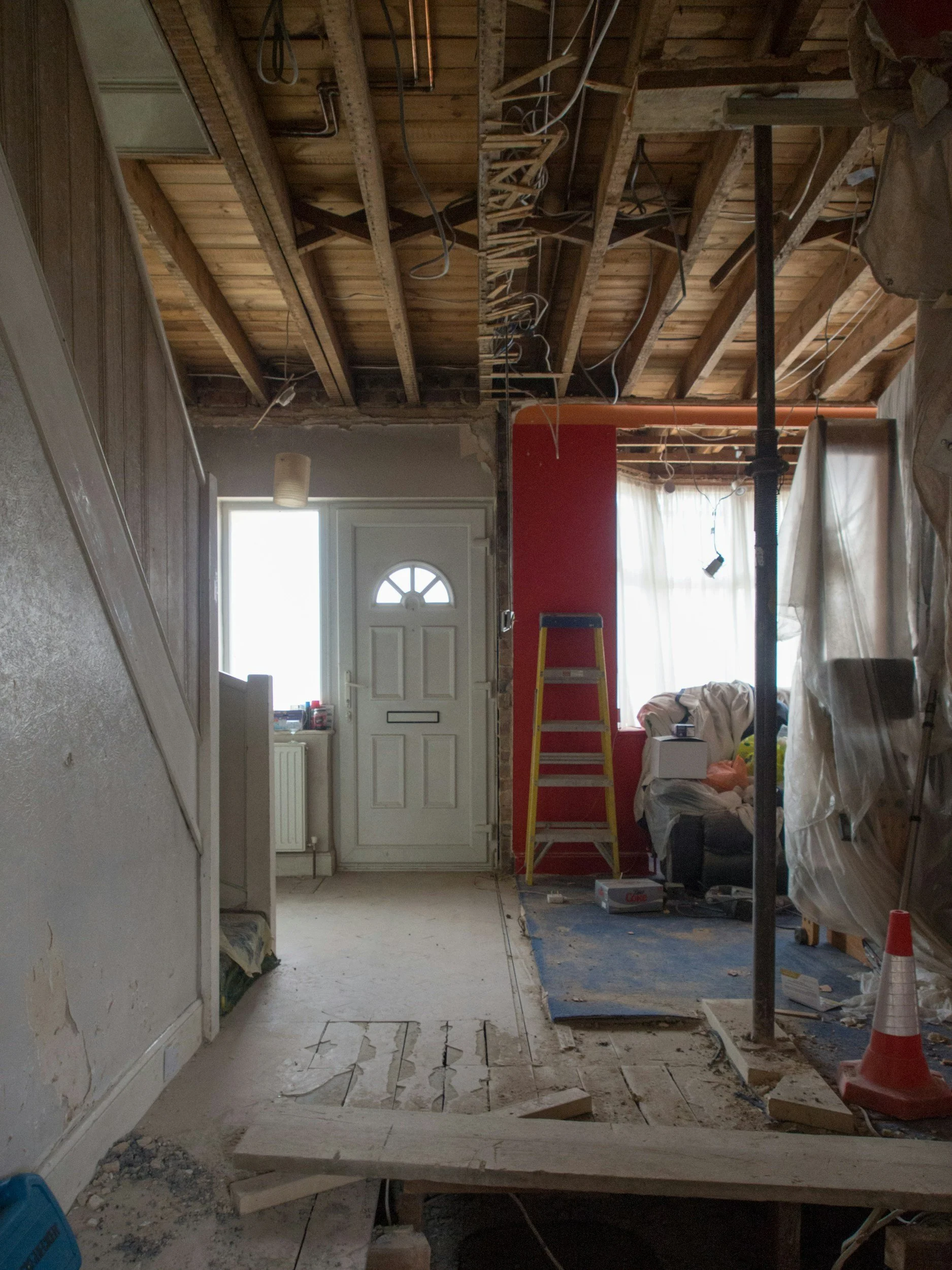 Interior of a house undergoing renovation, showing exposed ceiling beams, construction tools, and materials, with a partly removed floor and scattered debris.