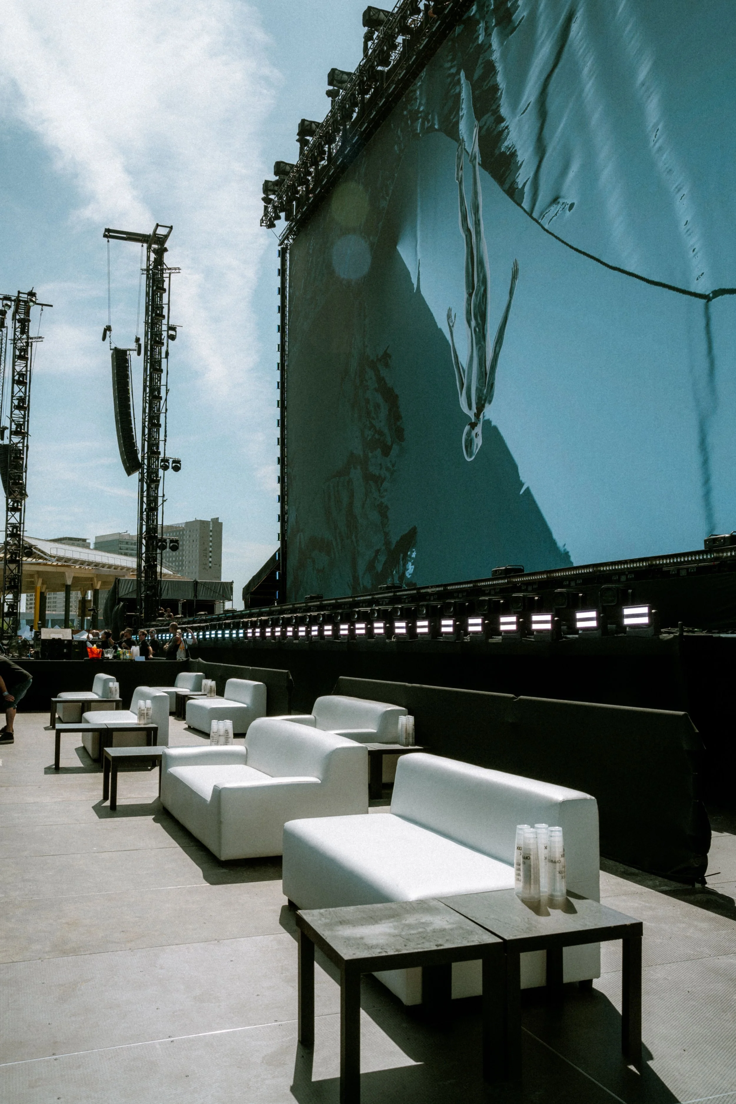 Empty white lounge chairs and small tables with water bottles on them on an outdoor stage with large screens in the background showing a water scene, and some construction or event equipment in the background during daytime.
