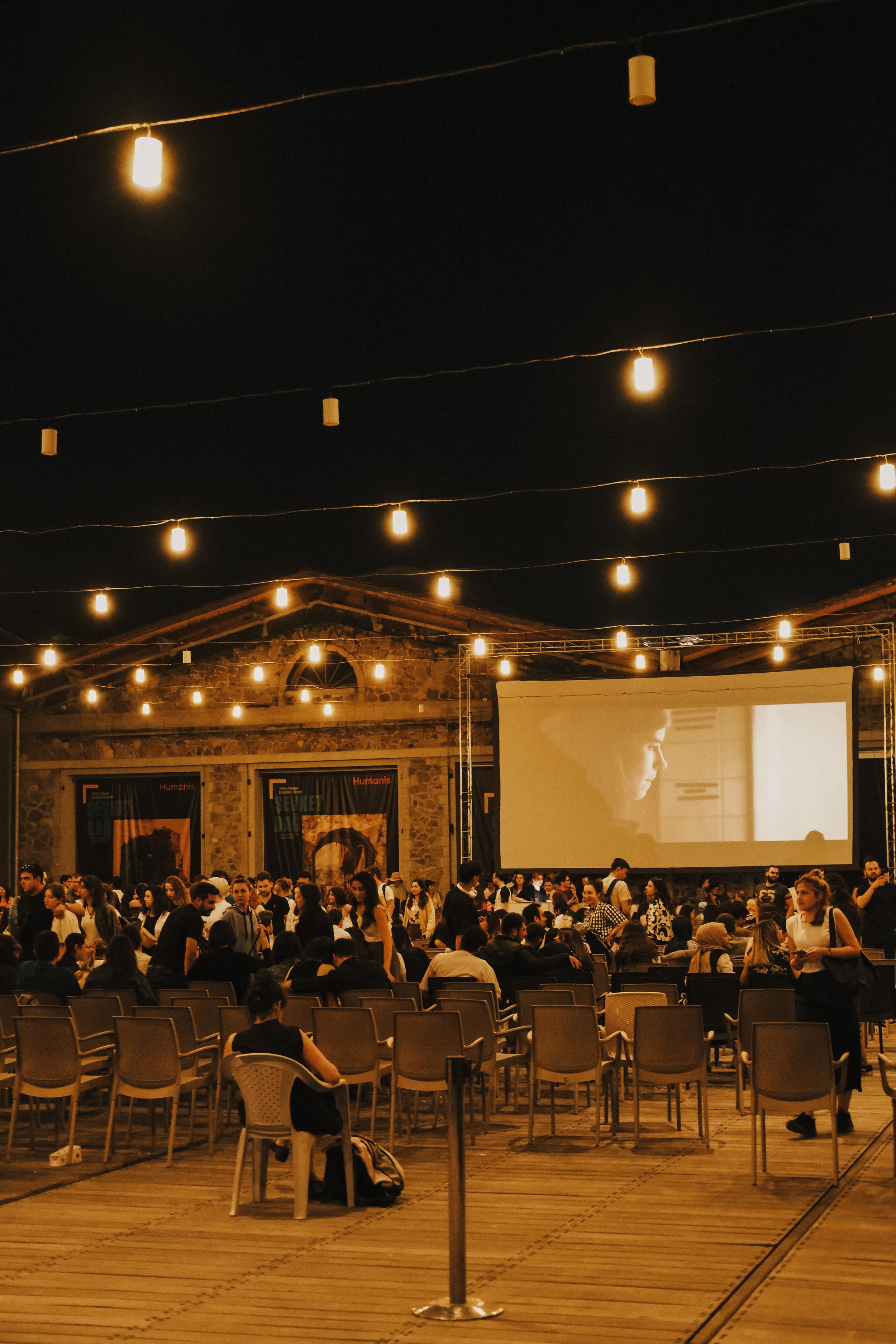 A crowded outdoor event at night with string lights overhead, an old brick building in the background, and people sitting and standing in front of a large projected screen showing a person's profile.