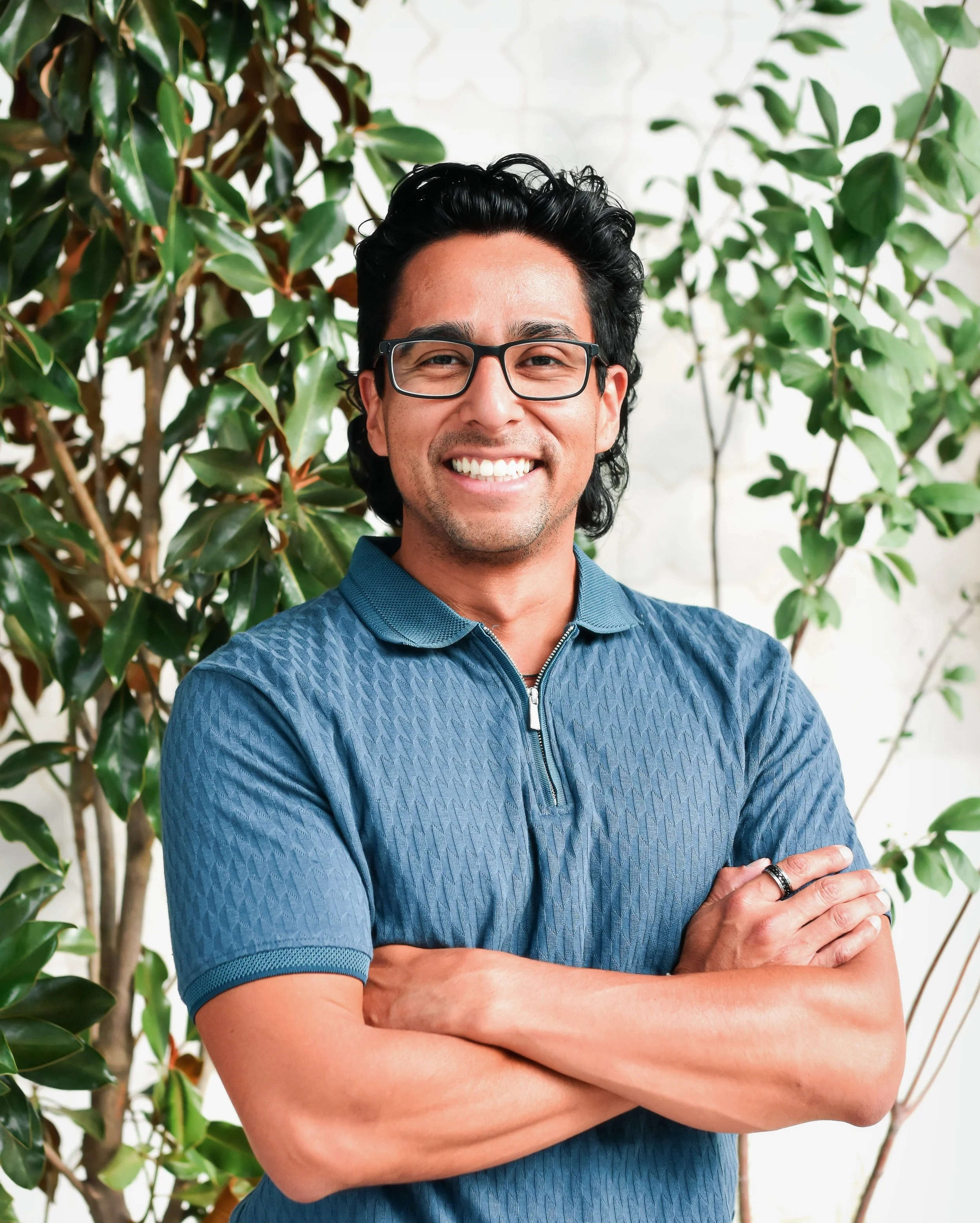 A smiling man with black hair, glasses, and a ring on his right hand, standing with arms crossed in front of green plants and a white wall.