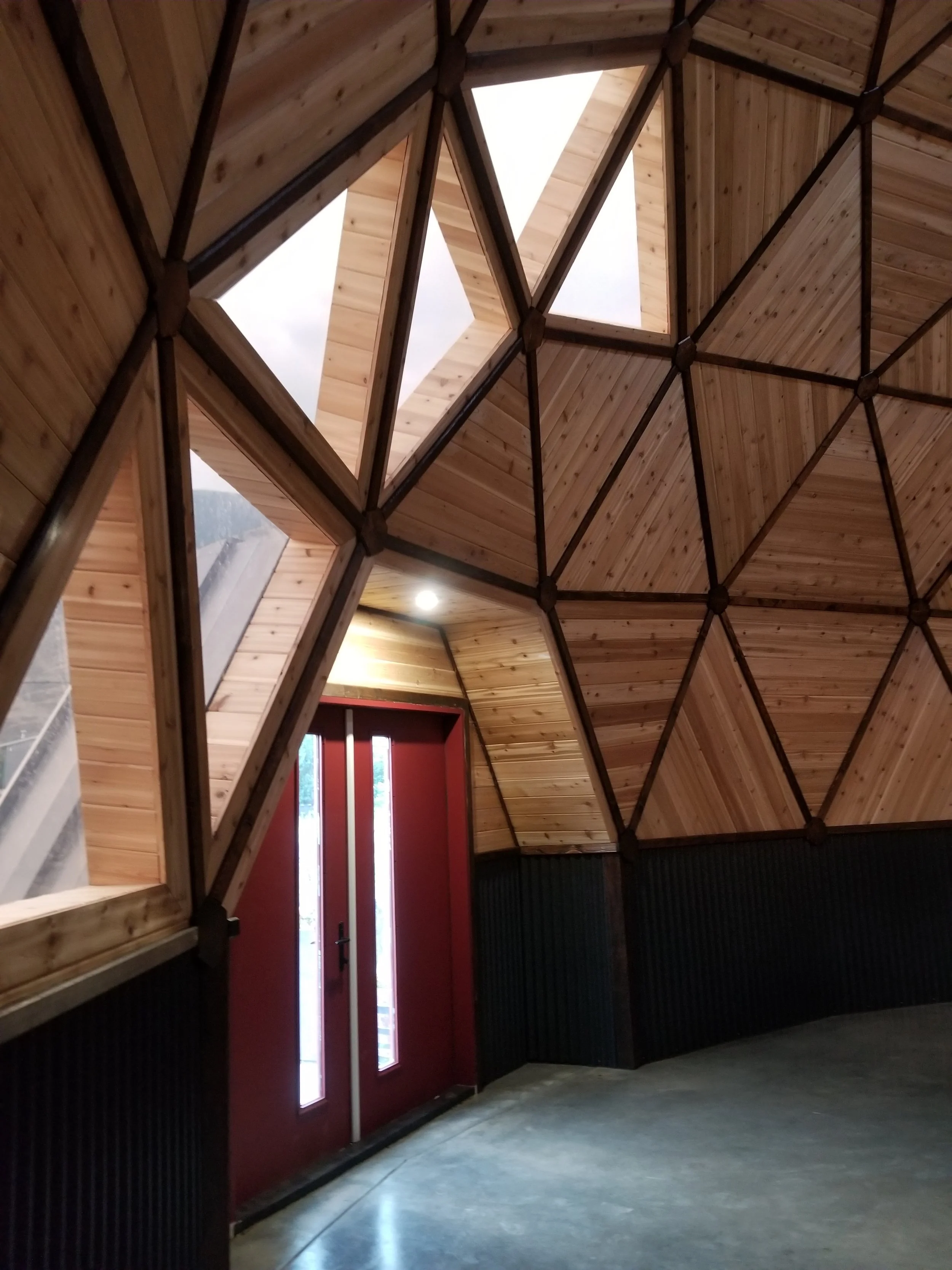 Interior view of a building with a high, wooden, geometric ceiling with window cutouts, a red door, and black wall paneling.