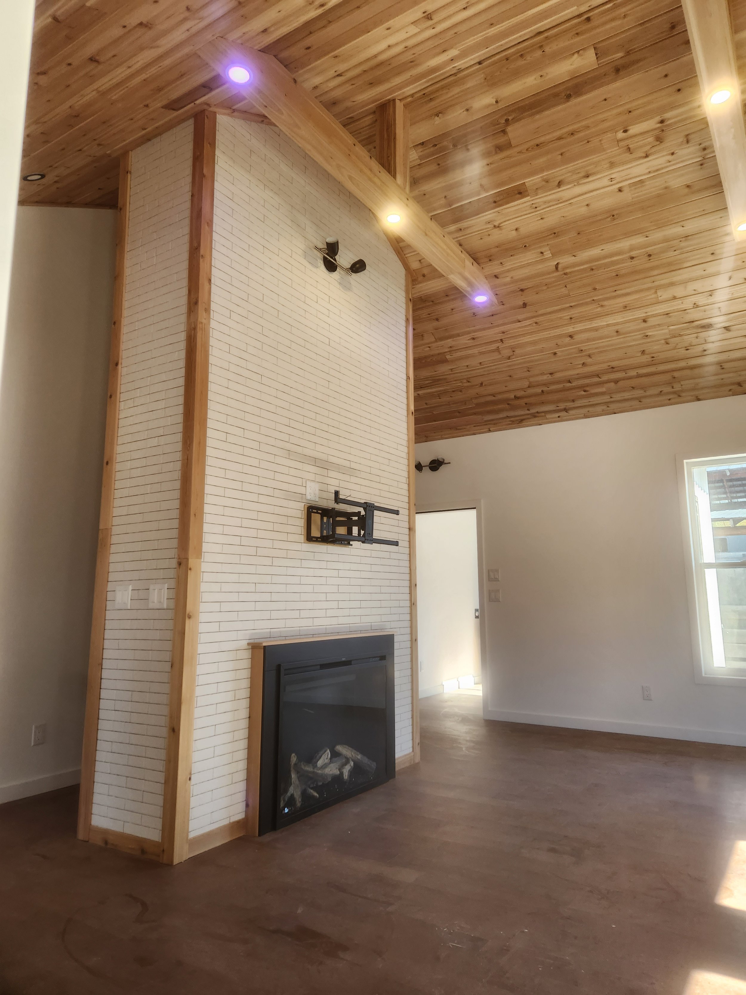 Interior of a room with a wood-paneled ceiling and a white brick fireplace wall. The ceiling has recessed lighting, and a wall-mounted light fixture is visible. There is a mountain-like black fireplace at the bottom of the wall. A window is on the right, and part of the wooden floor is visible.