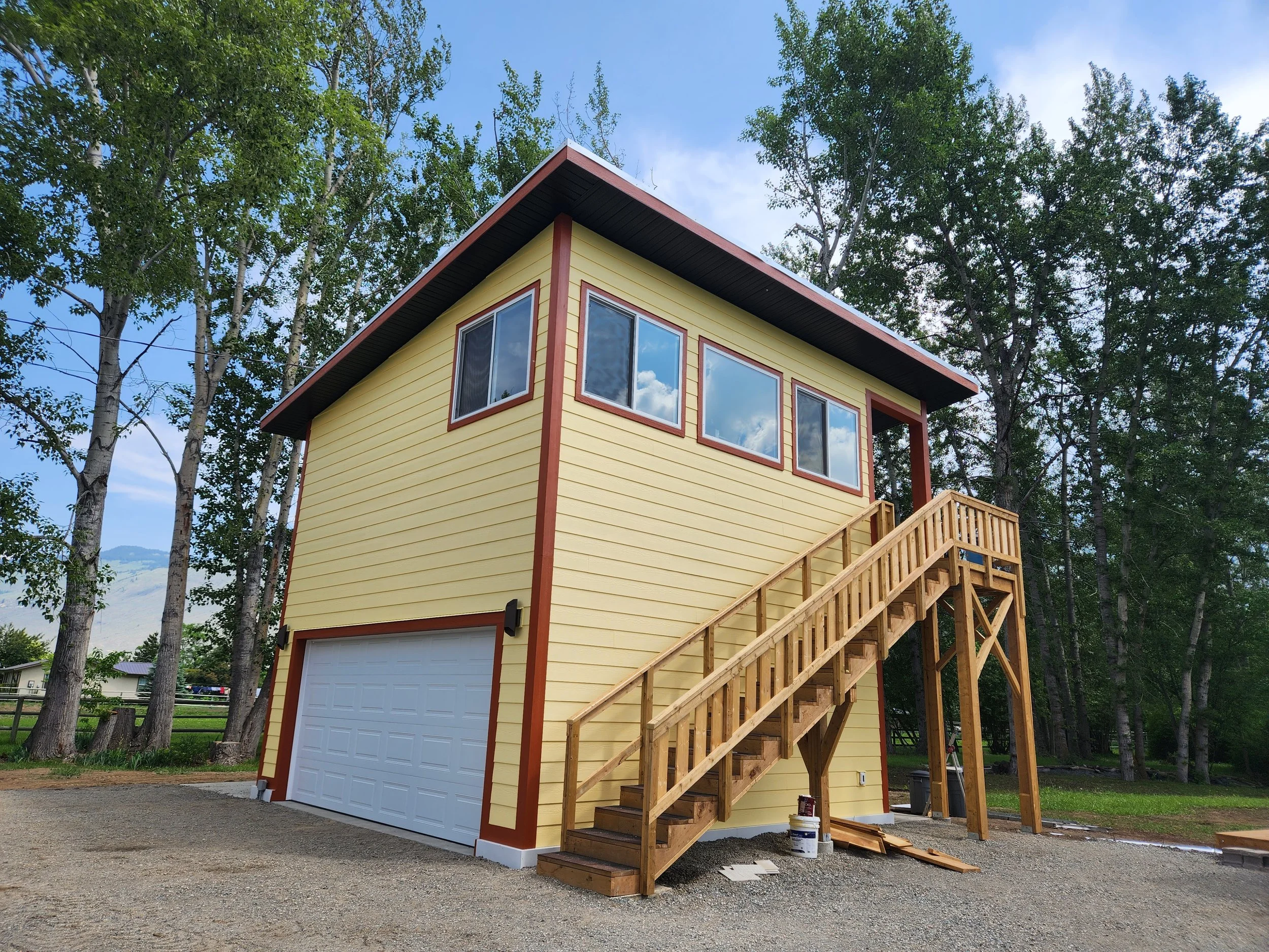 A yellow two-story garage with a white door, red trim, and wooden stairs leading up to an entrance, surrounded by tall trees under a partly cloudy sky.
