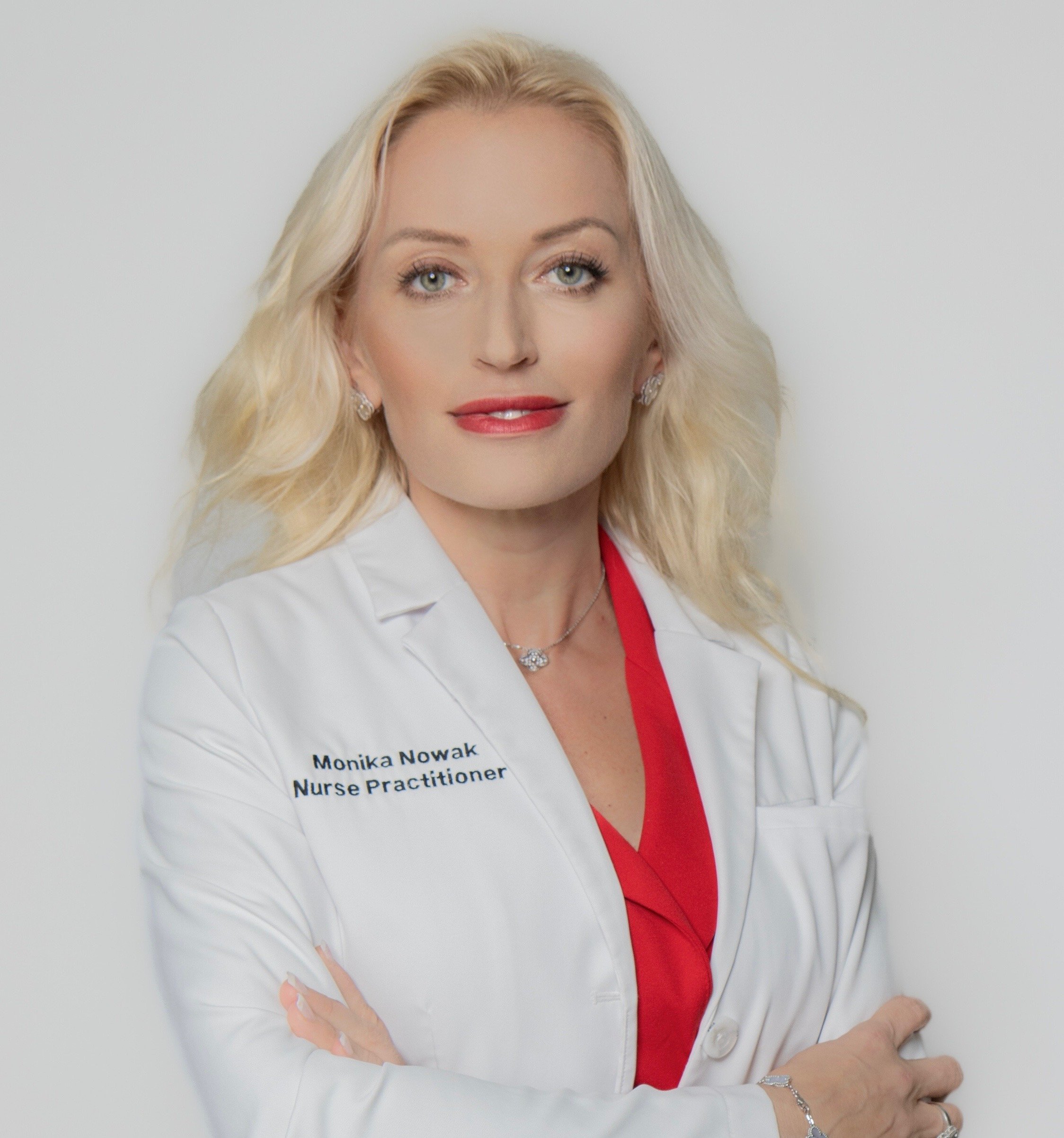 A woman with blonde hair, wearing earrings, a red top, and a white blazer with a name tag that reads 'Monika Nowak, Nurse Practitioner,' posing against a white background.