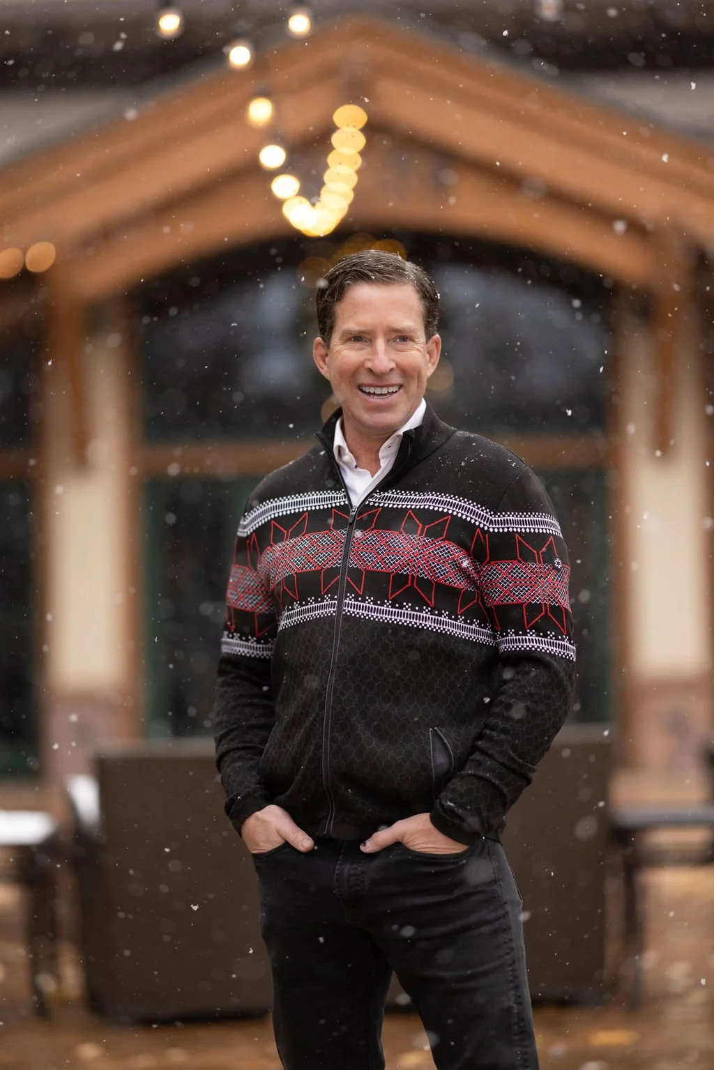 A smiling man wearing a black and red patterned Christmas sweater over a white shirt, standing outdoors in the snow at night, with blurred wooden structure and string lights in the background.