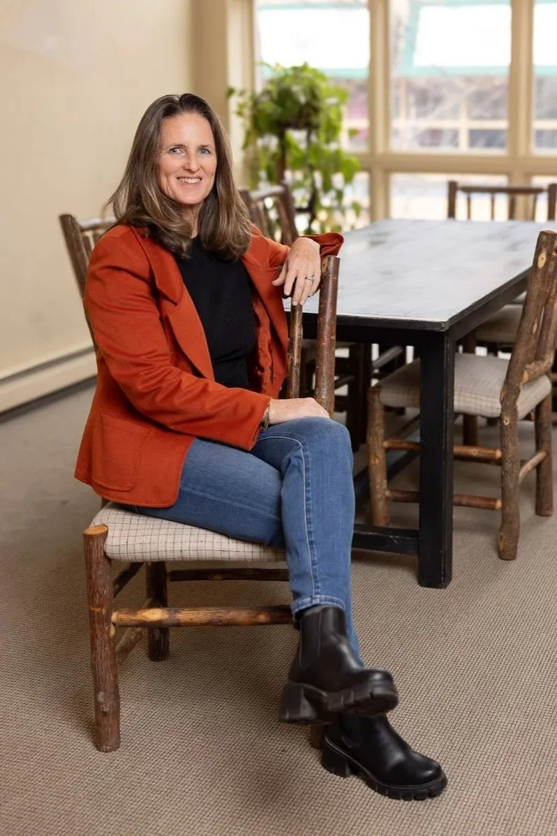 A woman with shoulder-length brown hair, smiling, wearing a black top, rust-colored blazer, blue jeans, and black boots, sitting on a wooden chair in a room with large windows, a black table, several wooden chairs, and a potted plant in the background.