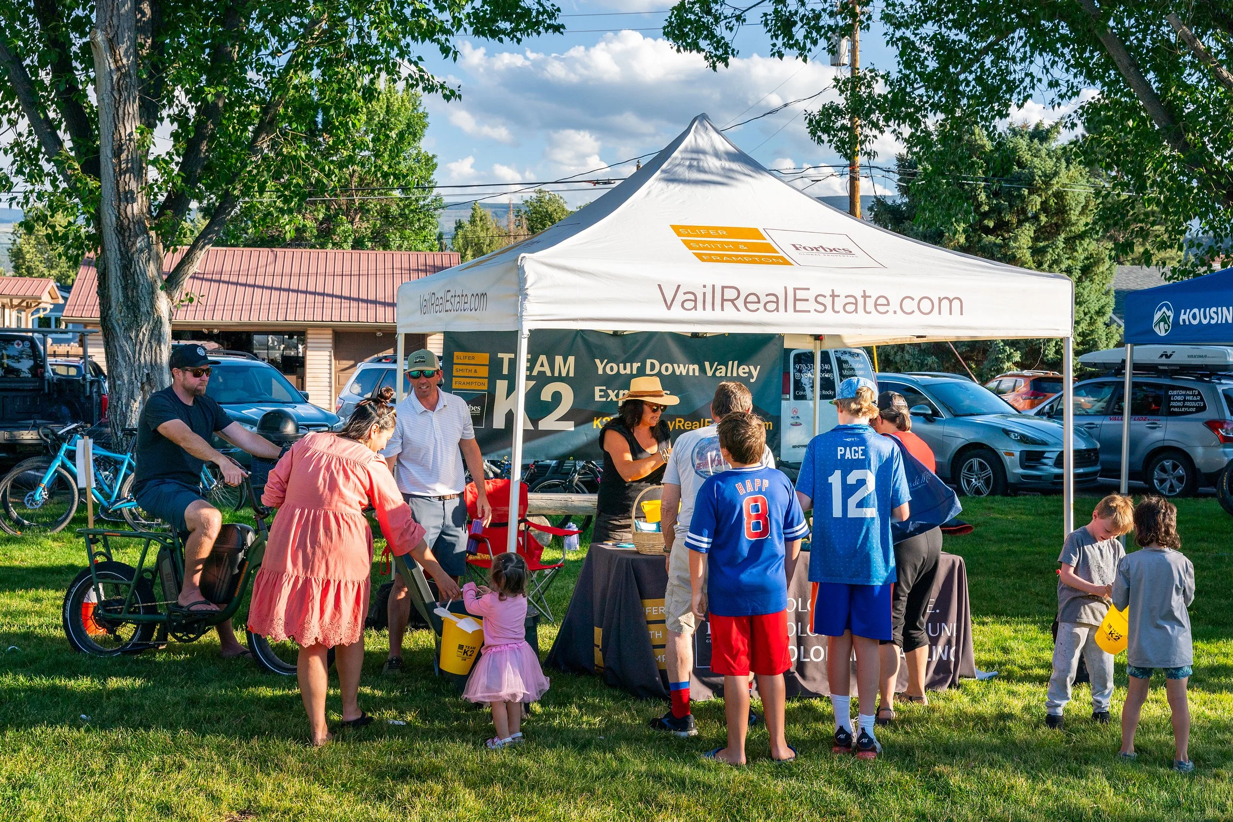People gathered at an outdoor event under a white tent with signs for VailRealEstate.com, called 'Your Down Valley Experts.' Several individuals are standing, sitting, or walking around, with some wearing sports jerseys, and children in the foreground. Parked cars, a bicycle, and trees are visible in the background.