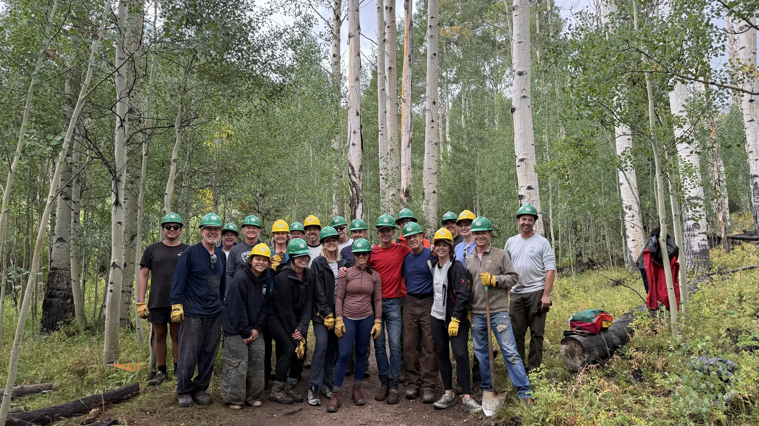 Group of people wearing helmets and gloves, standing together in a forested area with tall trees and green foliage.