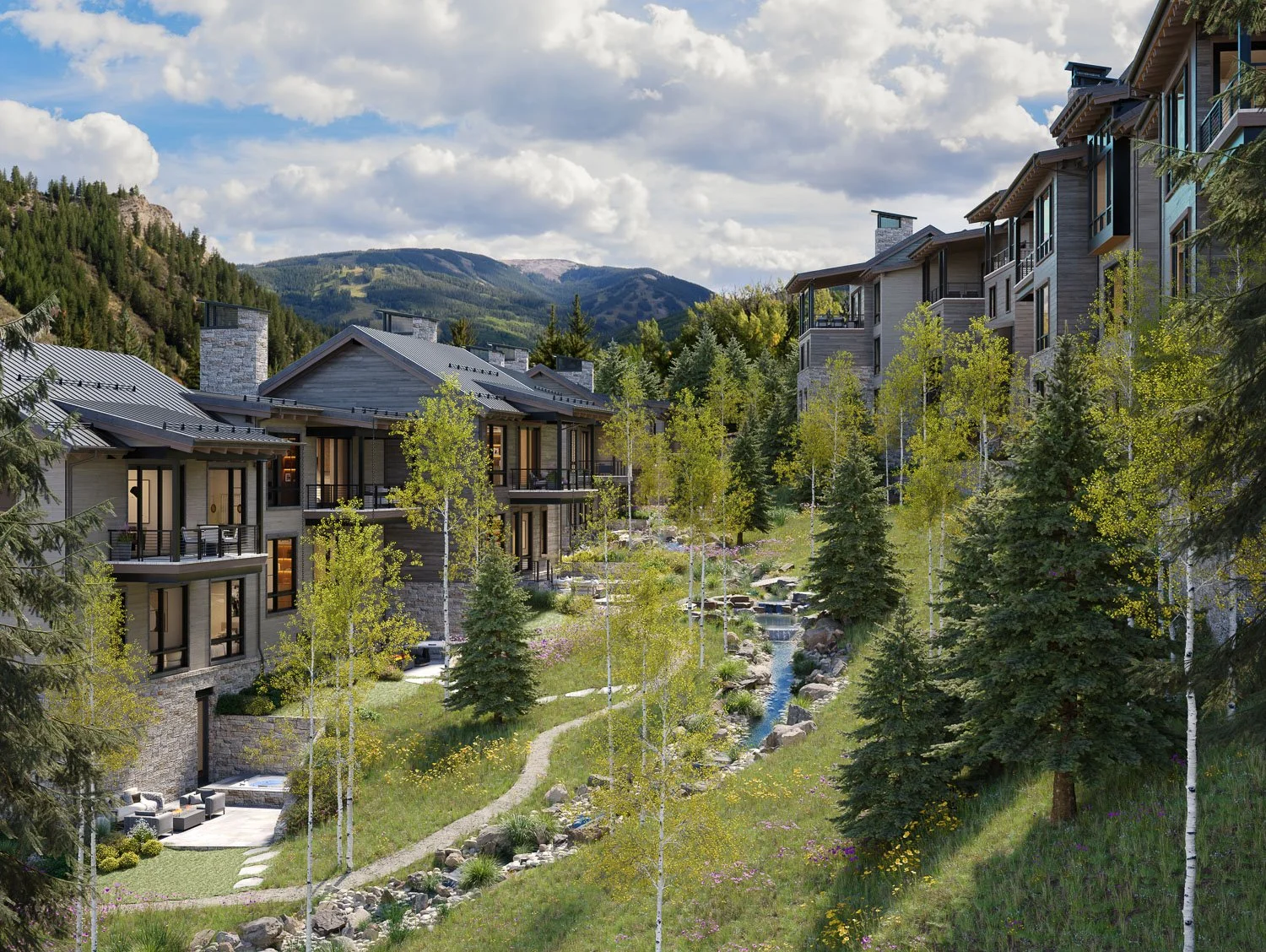 A scenic view of modern residential buildings nestled in a lush, green mountain landscape with a small stream running through the grassy area.