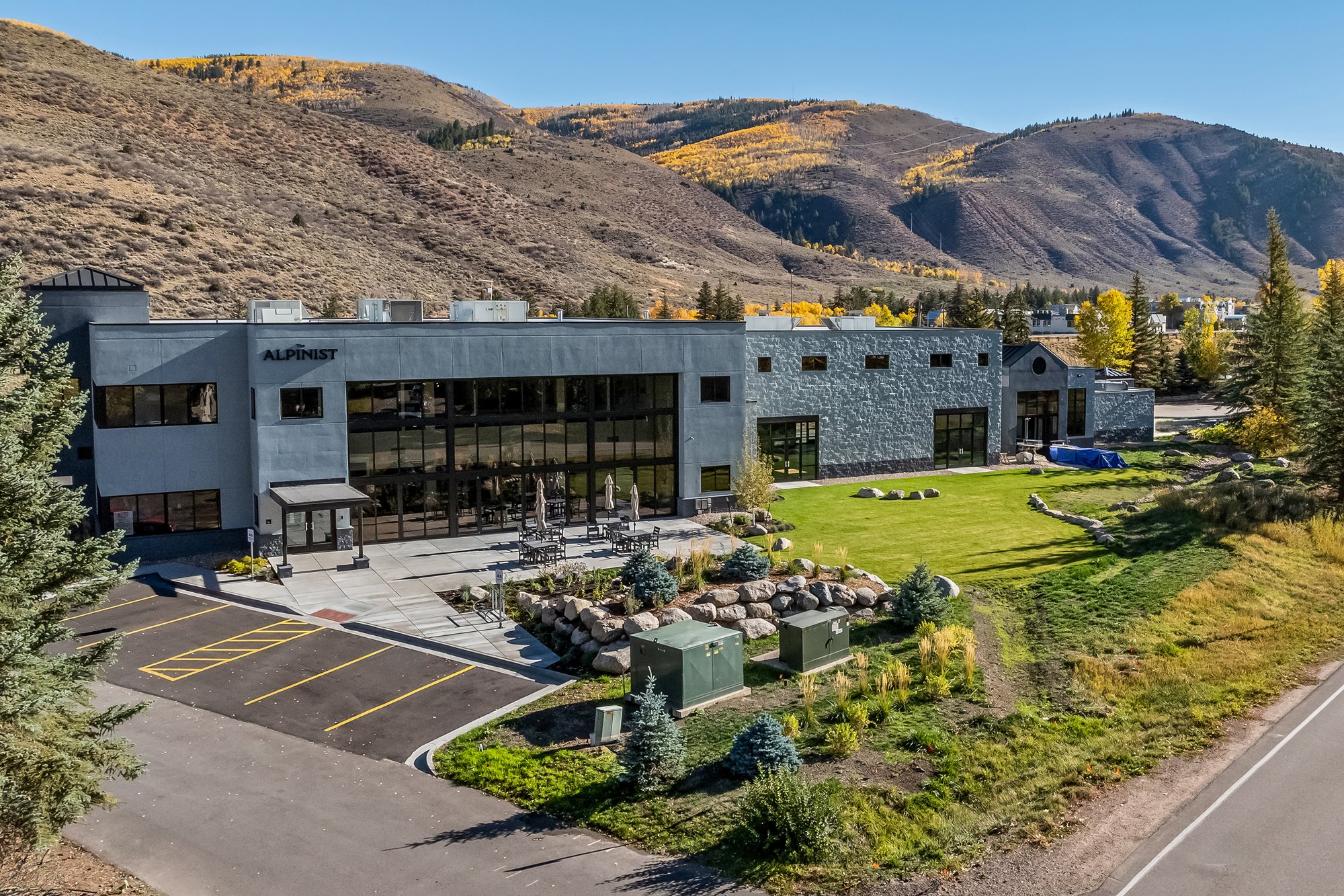 Office building named 'The Alpinist' with outdoor seating area, surrounded by trees, with mountains in the background and a clear blue sky.