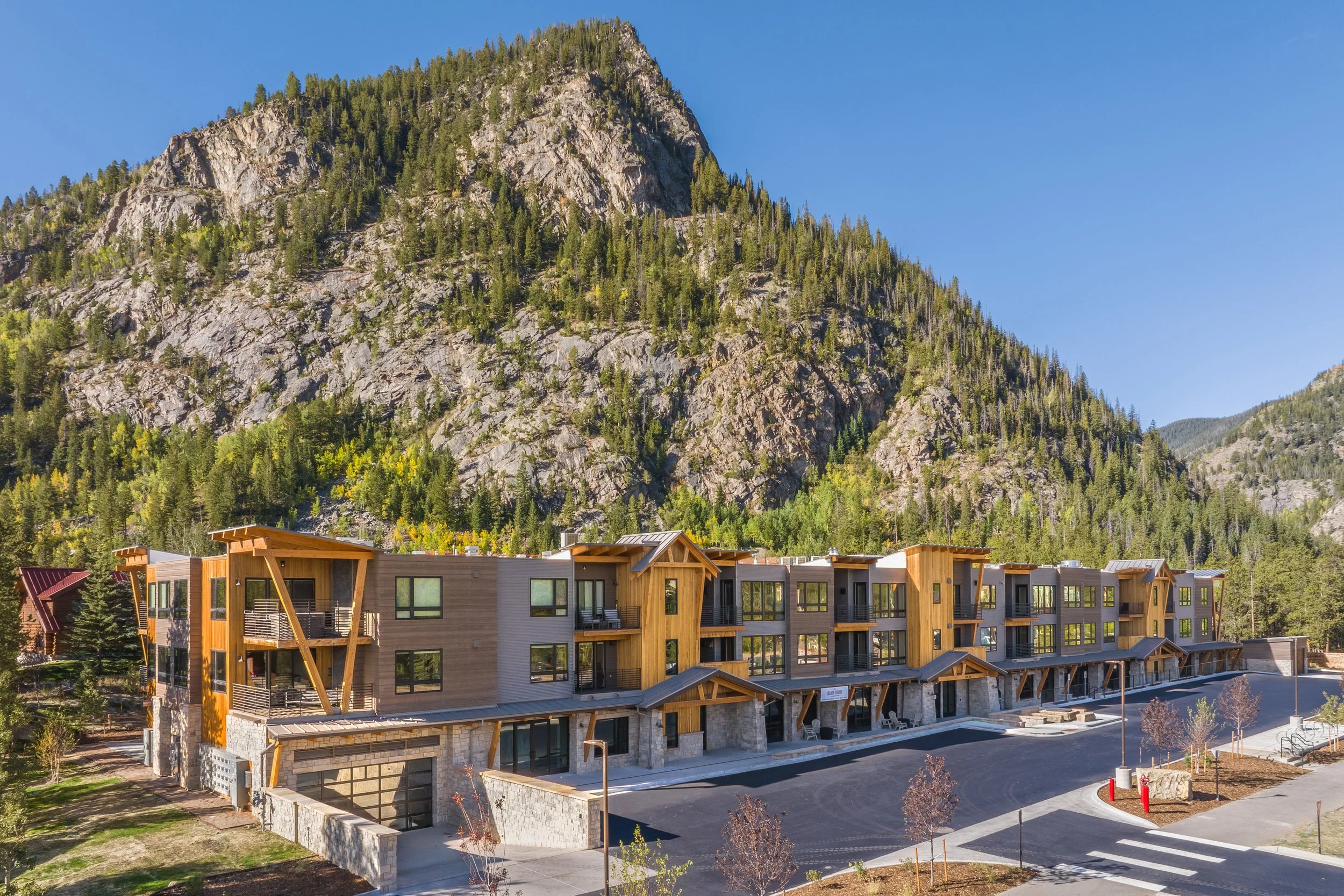 Modern residential buildings with wooden and stone exterior in front of a forested mountain landscape under a clear blue sky.