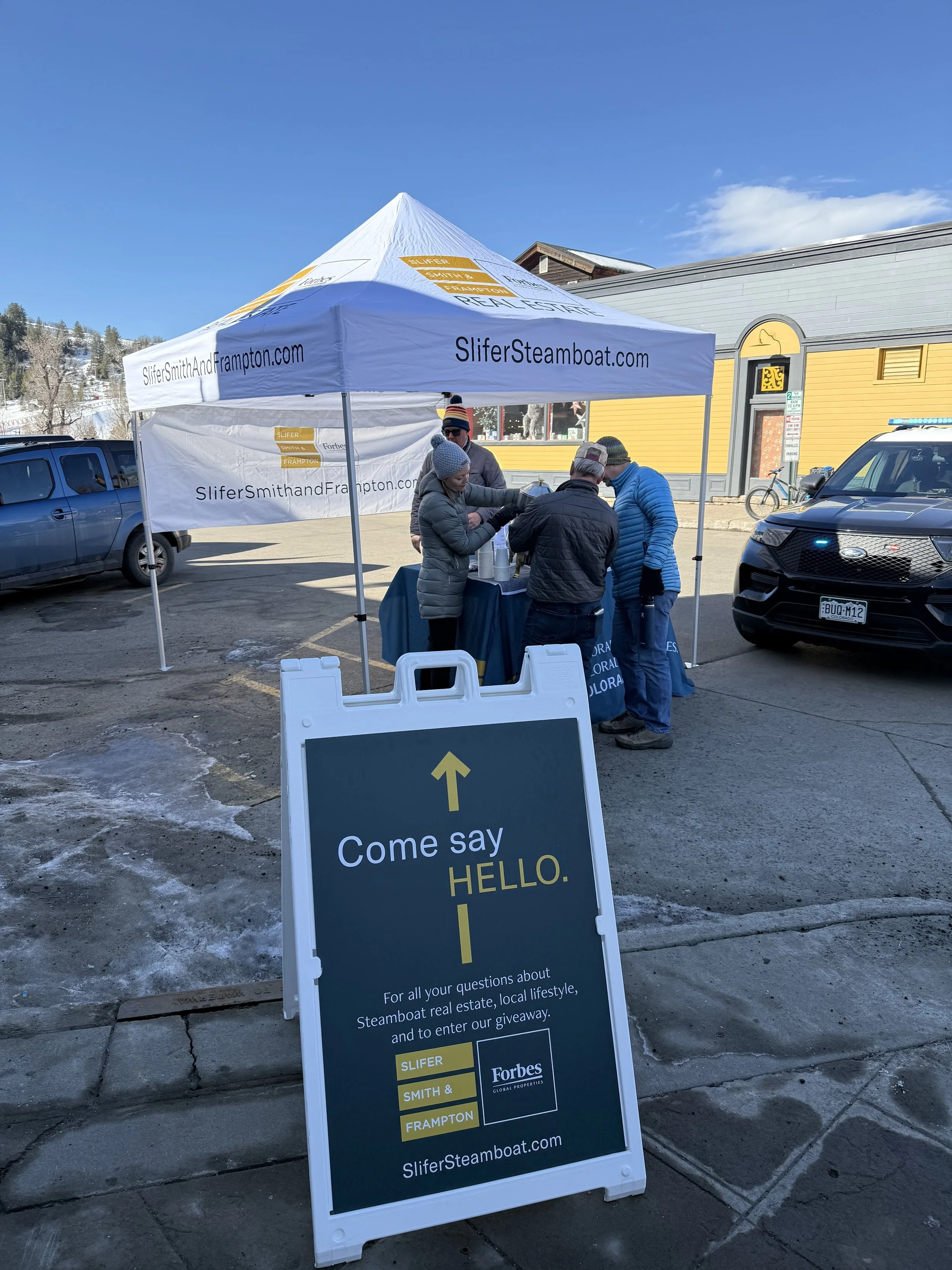 A white sandwich board sign with yellow and white text, featuring an arrow pointing upward, placed outdoors on a paved surface. The sign invites viewers to come say hello and provides information about a steamboat-related event or giveaway, with addi