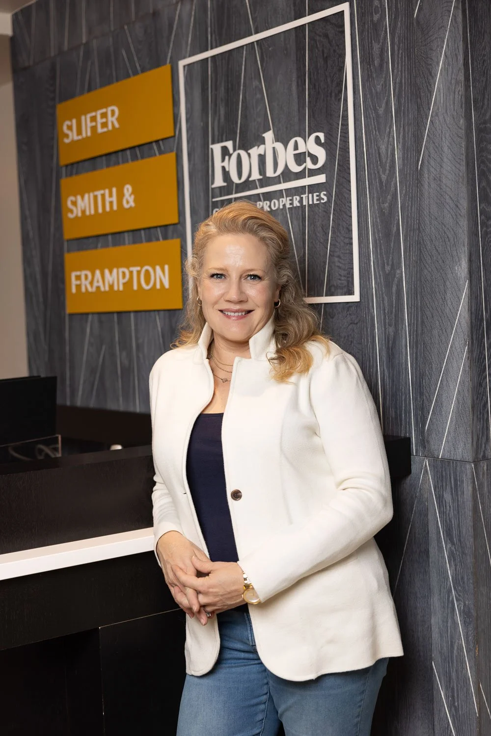 A woman with blonde hair, wearing a white jacket over a dark top and jeans, posing in front of a wall with Forbes and real estate agency signs.
