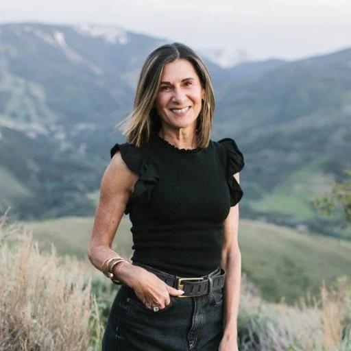 A woman standing outdoors in front of mountains and greenery, smiling at the camera.
