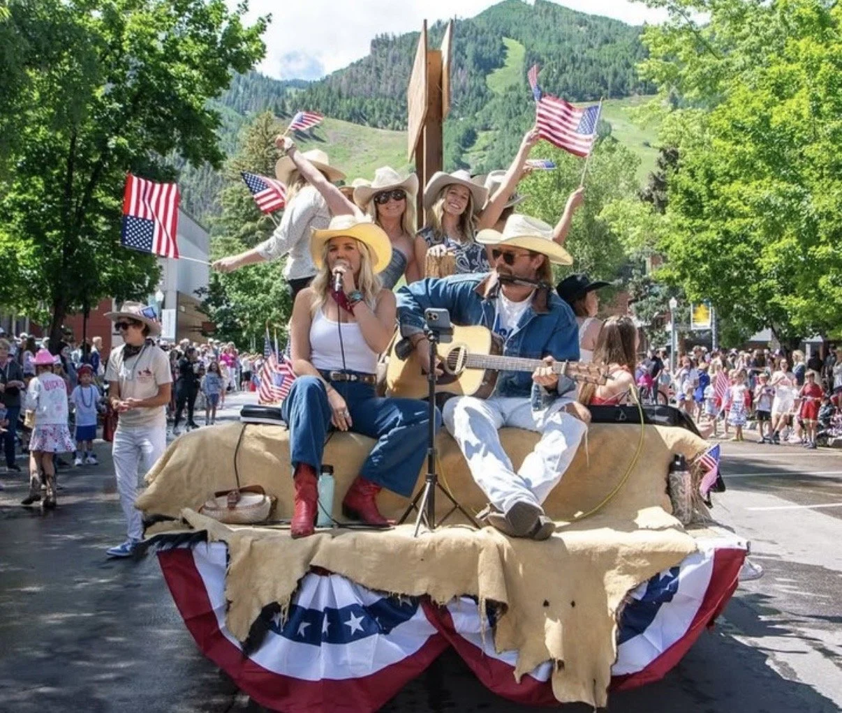 A parade float with people dressed in western attire, playing instruments and waving American flags, surrounded by a crowd of spectators in a mountainous town.