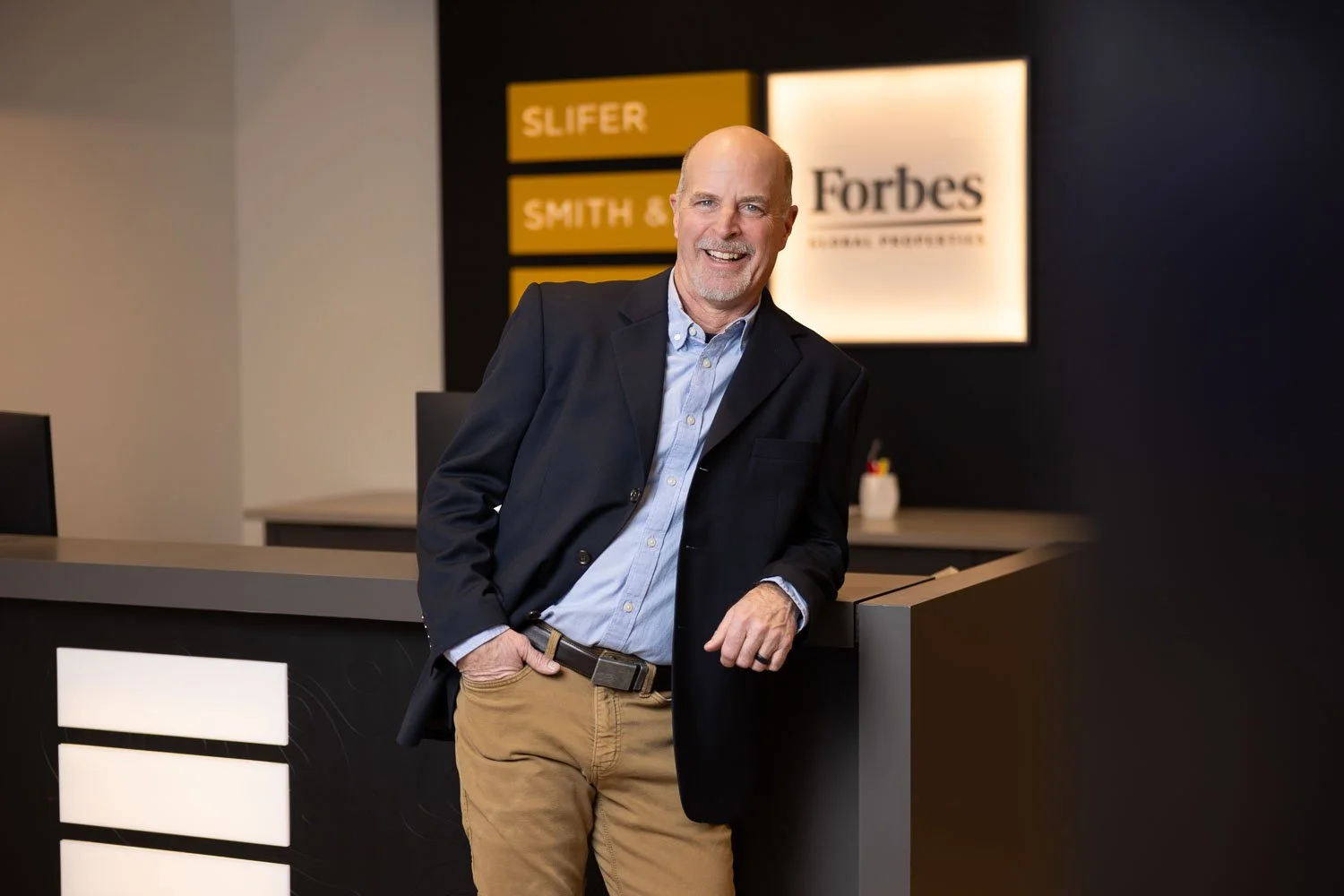 A man in a dark blazer and khaki pants leaning on a modern reception desk inside an office building.
