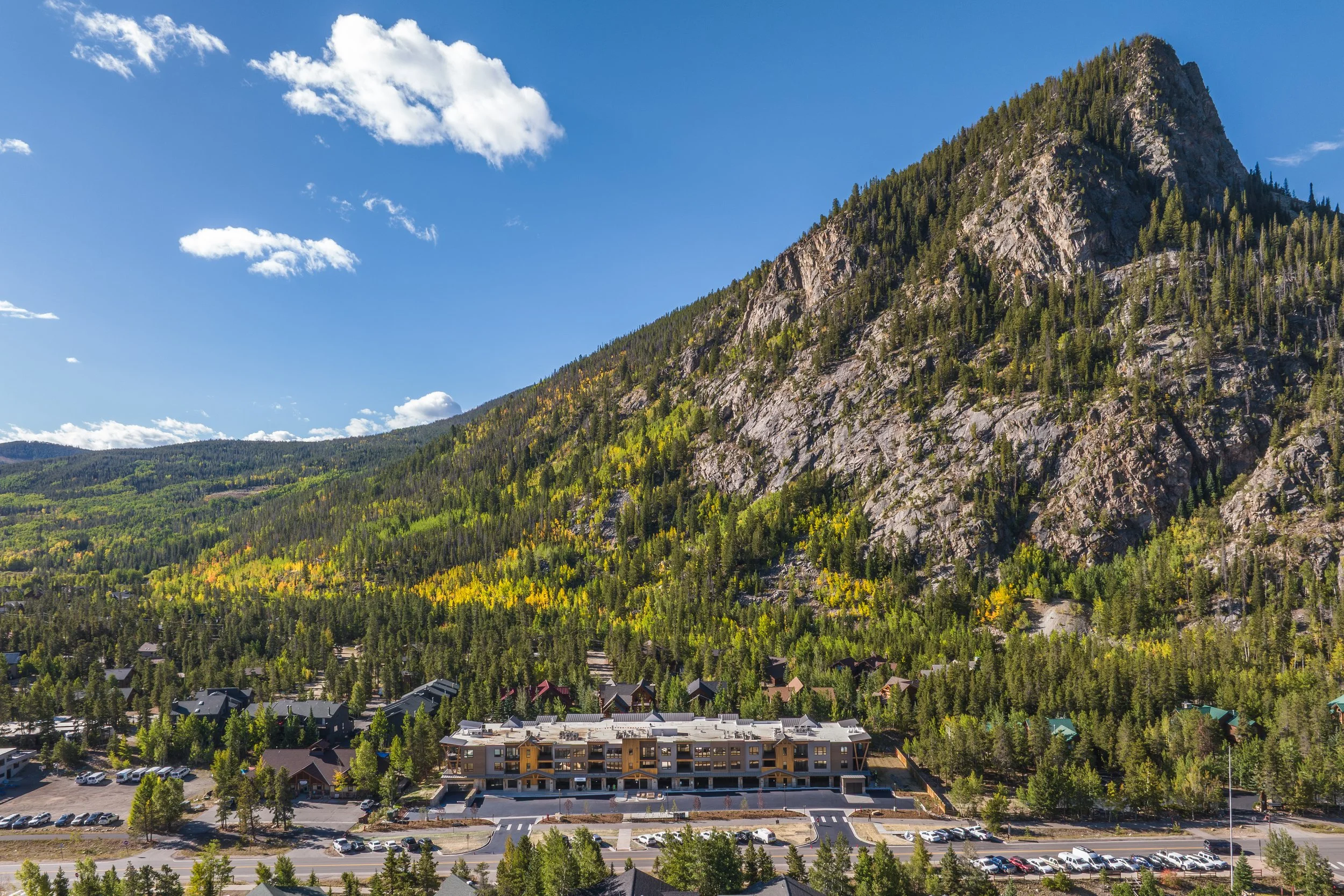 A mountain with a forested slope and small town buildings at the base, under a blue sky with scattered clouds.