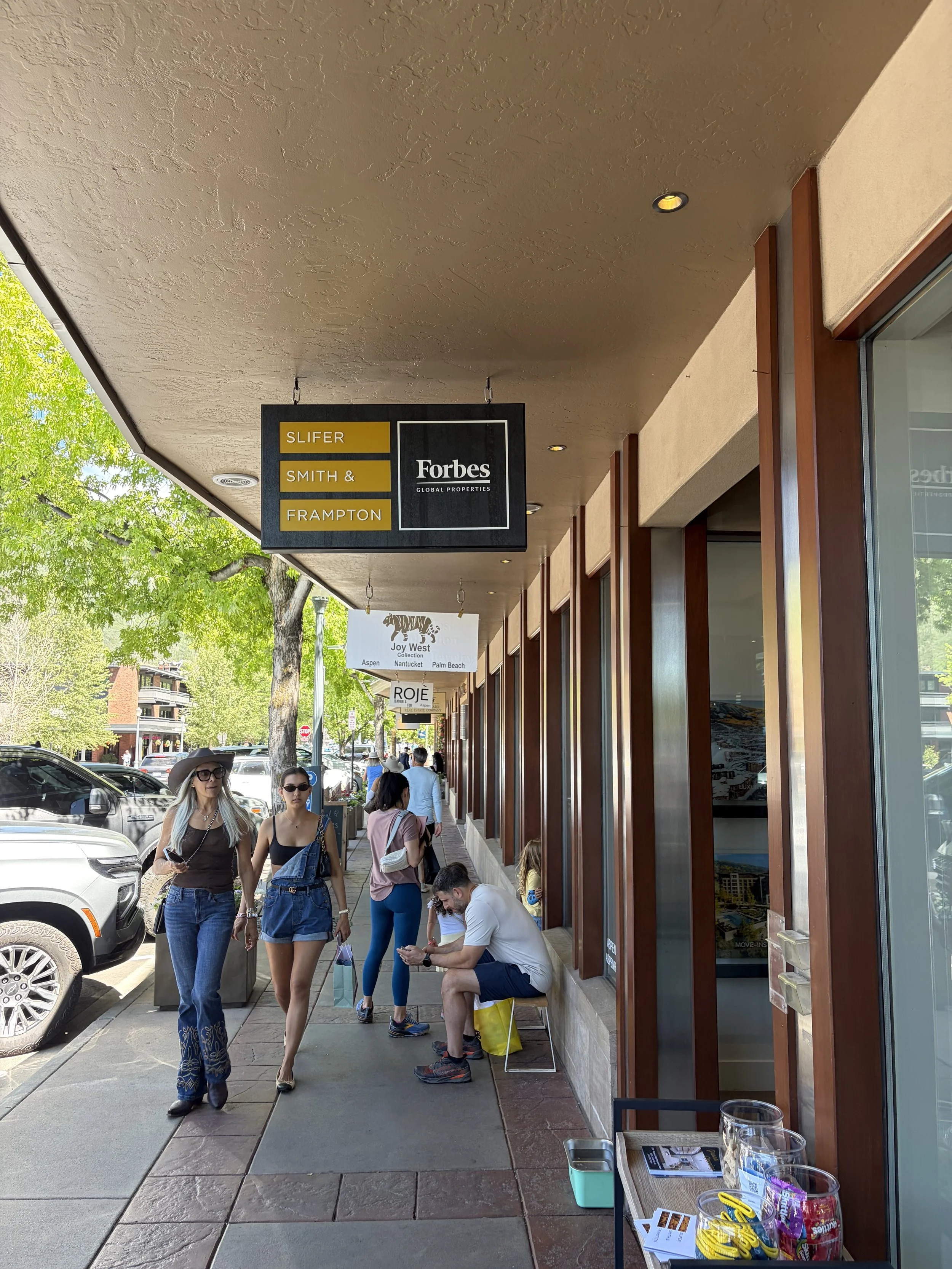 Sidewalk scene with pedestrians, storefronts, and hanging signs in a shopping area, sunny day with trees and parked cars.