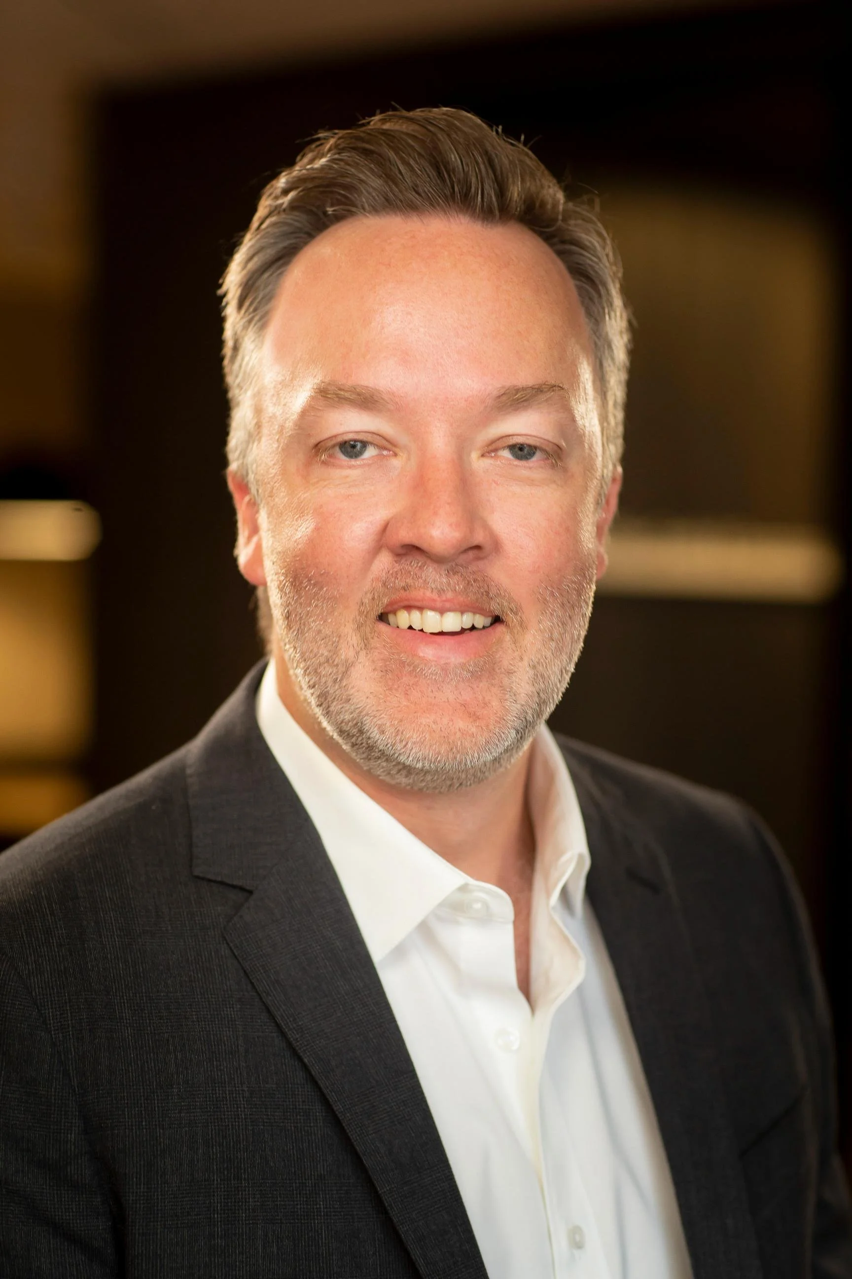 A middle-aged man with gray hair and a beard, wearing a dark suit and white shirt, smiling in a professional setting.