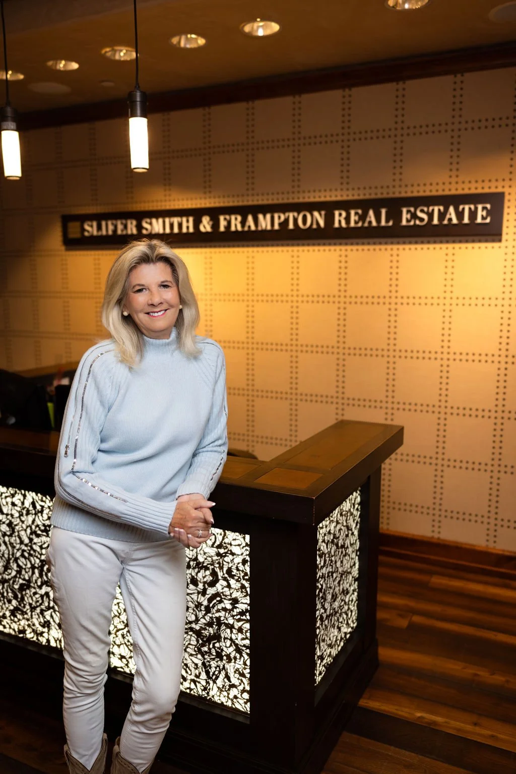 A smiling woman in a light blue sweater and light-colored pants standing in front of a reception desk inside a real estate office with a sign that reads 'Slifer Smith & Frampton Real Estate'.