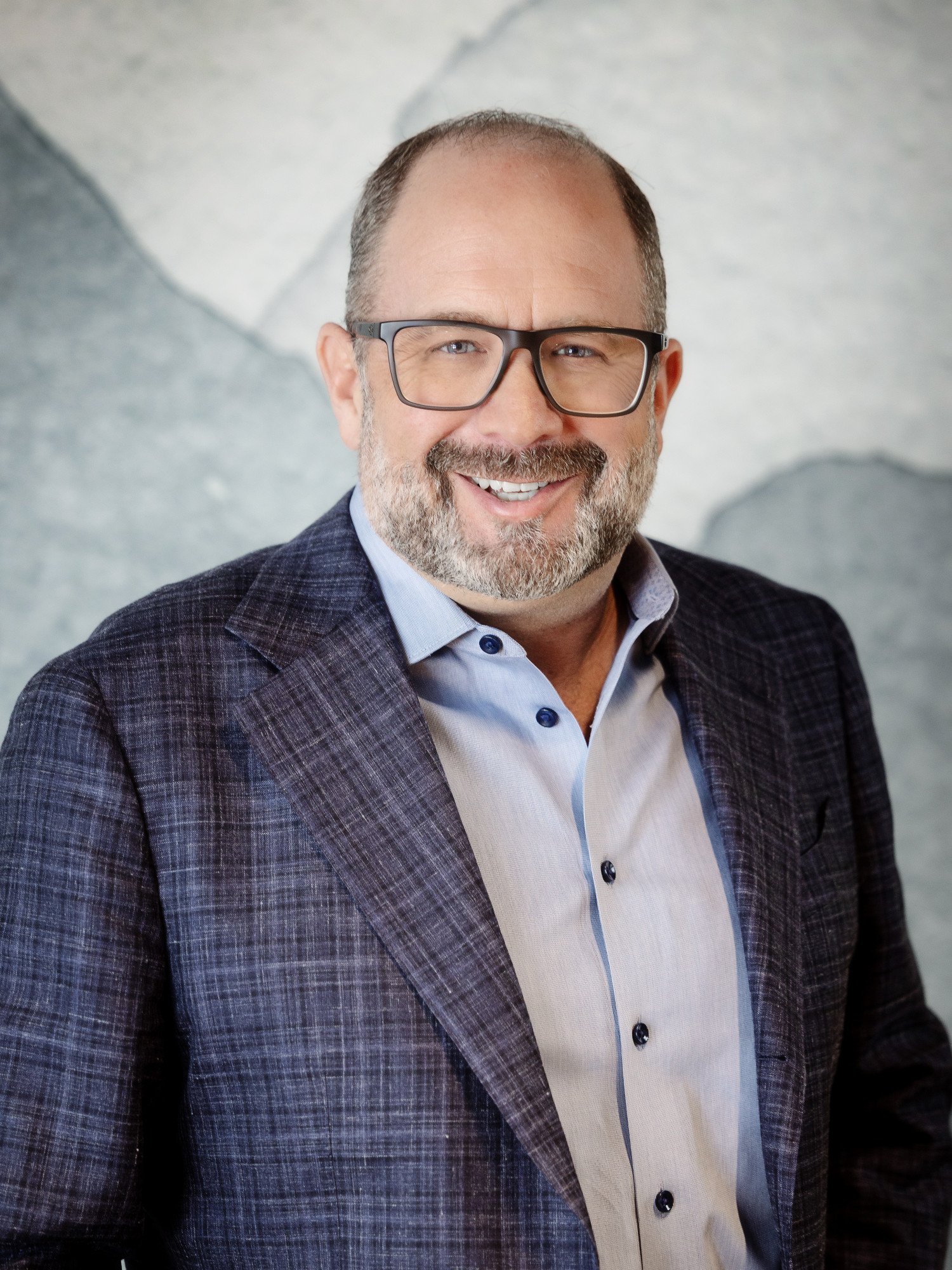 A smiling man with glasses, a beard, and short hair dressed in a light-colored shirt and a dark plaid blazer standing against a light gray stone wall.