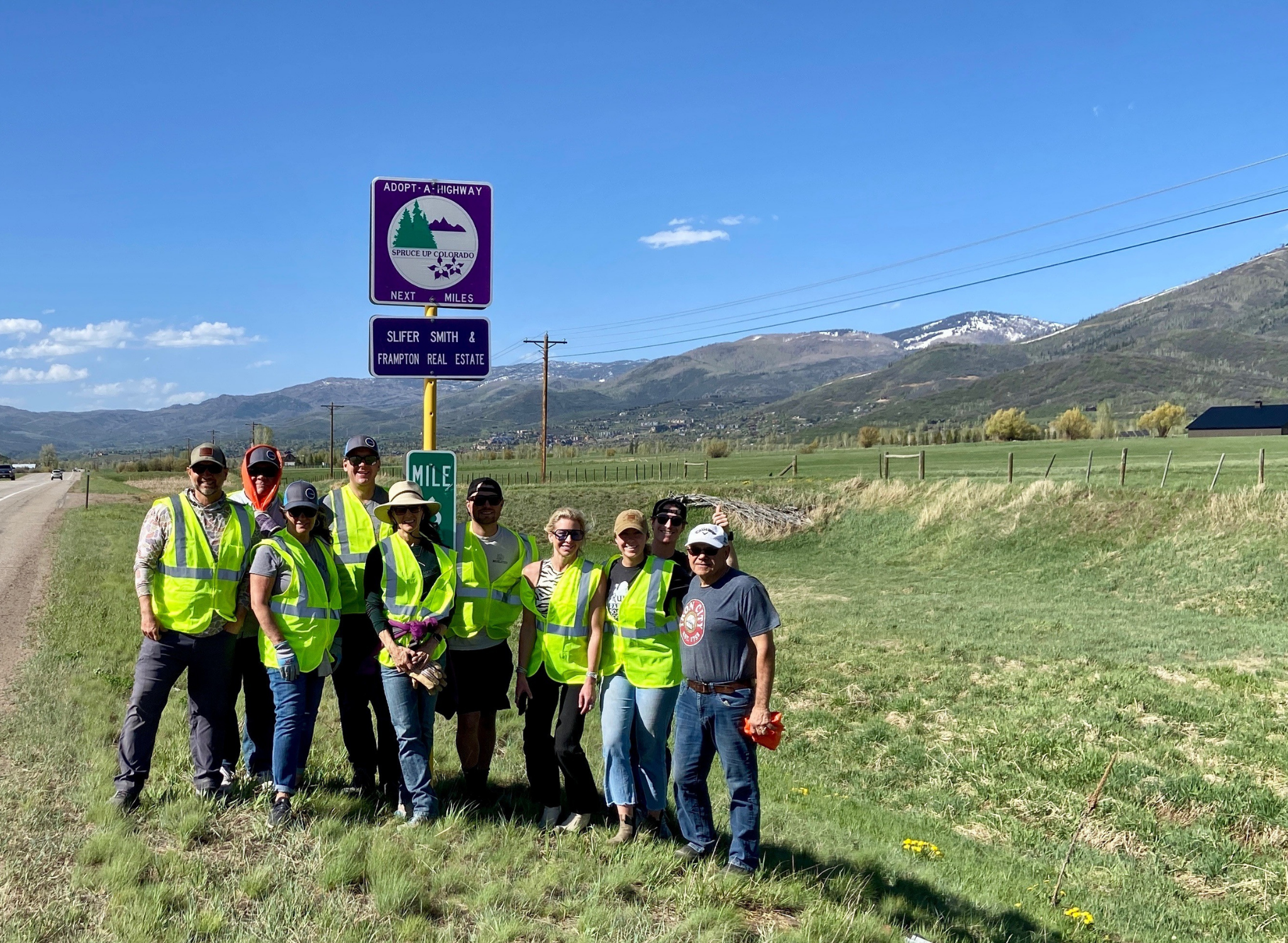 A group of people wearing yellow reflective vests standing outdoors on a grassy area with mountains in the background, a blue sky with some clouds, and highway signs that say 'Adopt-A-Highway' and 'Next Miles'.