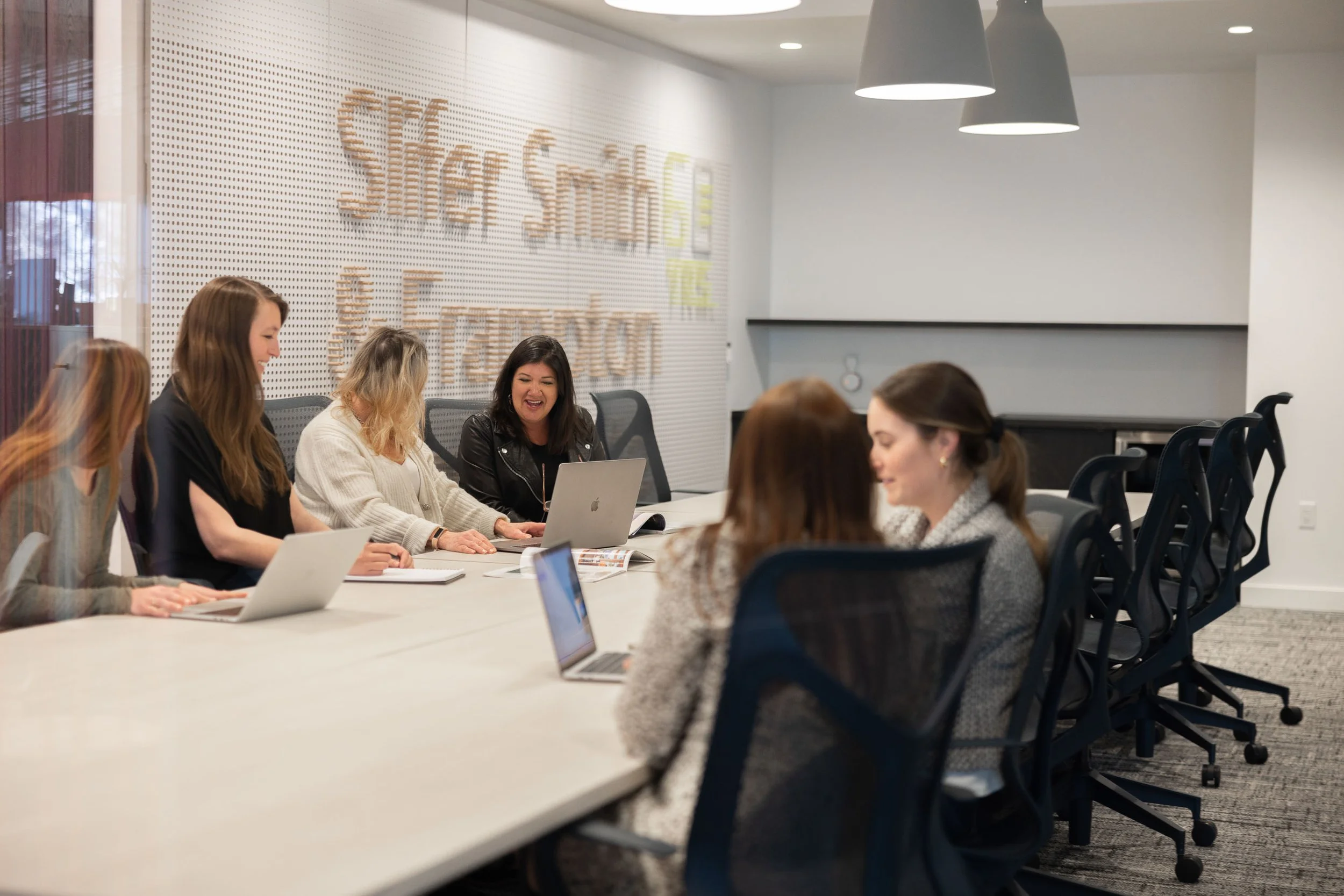 A group of women sitting at a conference table in a modern office, engaged in a discussion with laptops and notebooks open.