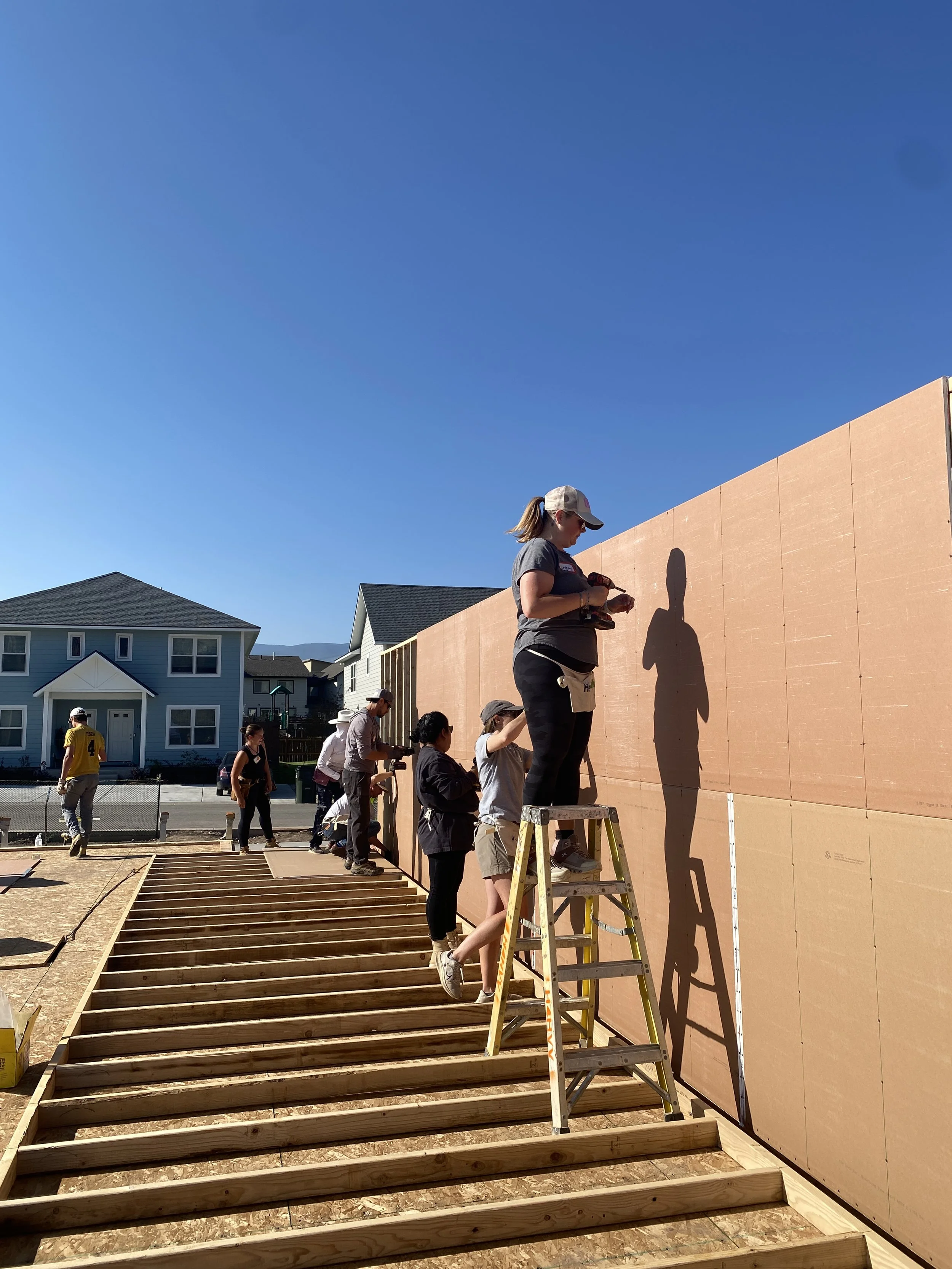 People working together building a wooden staircase and wall on a construction site under a clear blue sky in a suburban neighborhood.