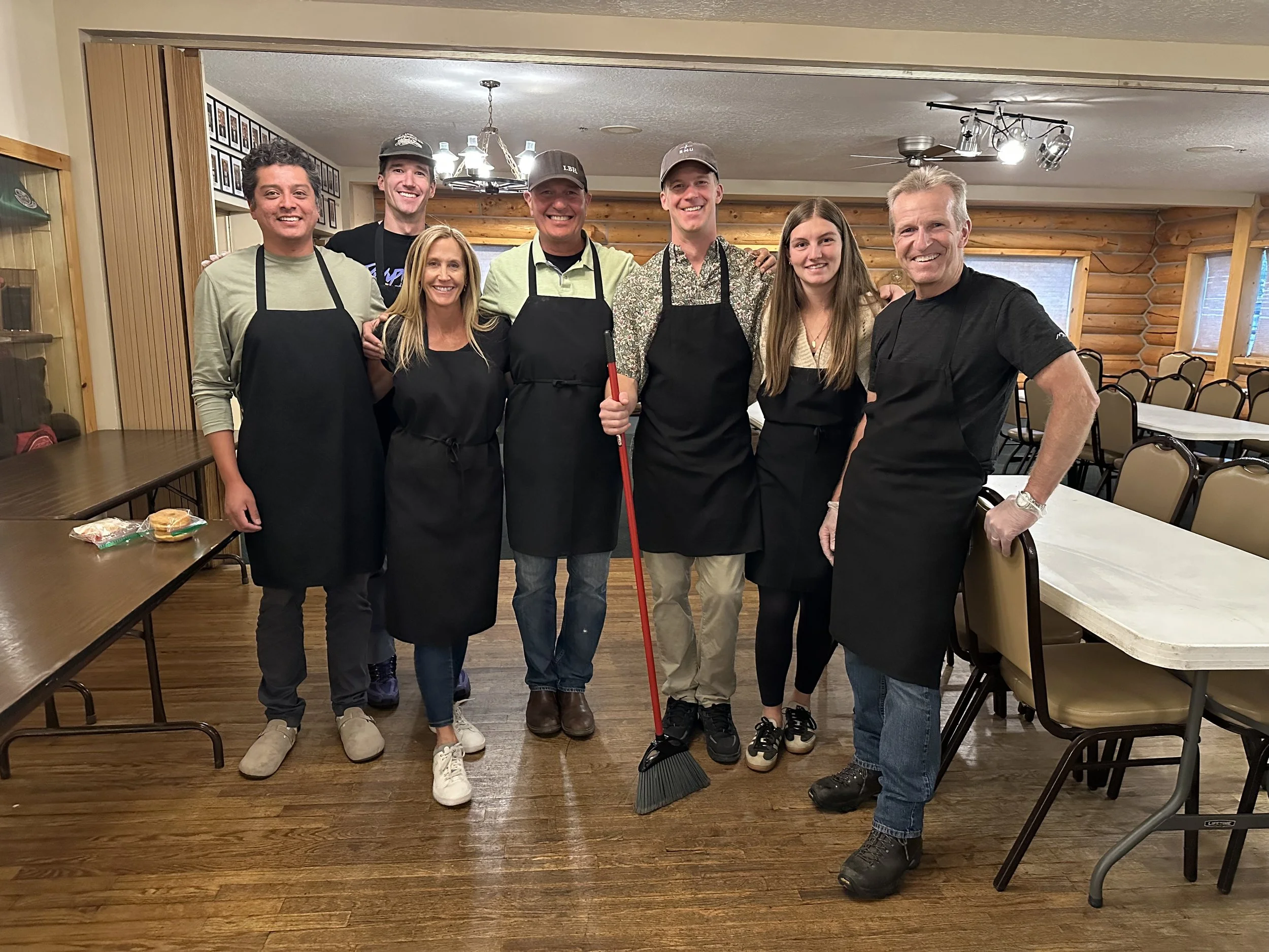 Group of seven people standing close together in a restaurant, smiling at the camera. They are wearing black aprons, and one person is holding a broom. The background includes wooden walls, tables, chairs, and framed photos on the wall.