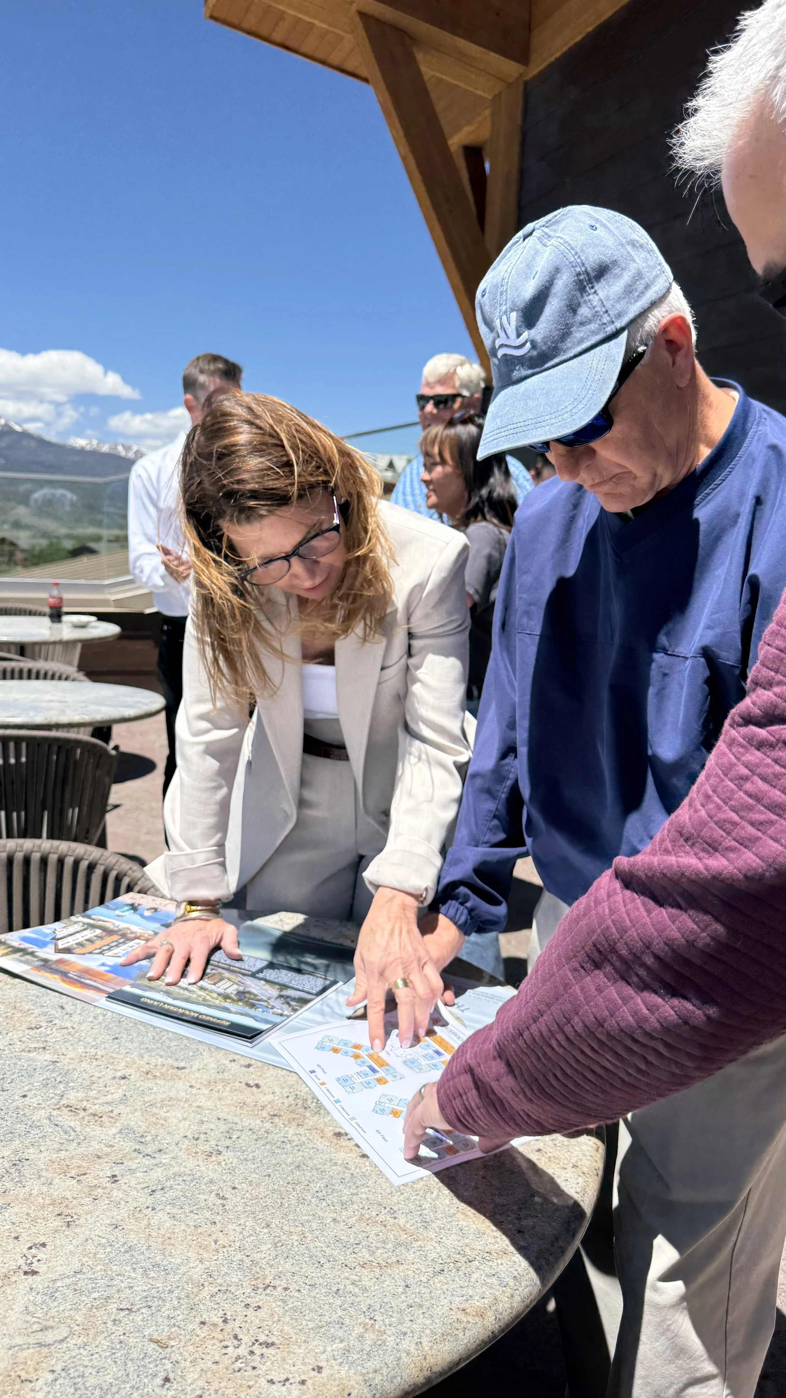 Group of people gathered around a table, looking at maps or brochures outdoors on a sunny day, with mountains and blue sky in the background.