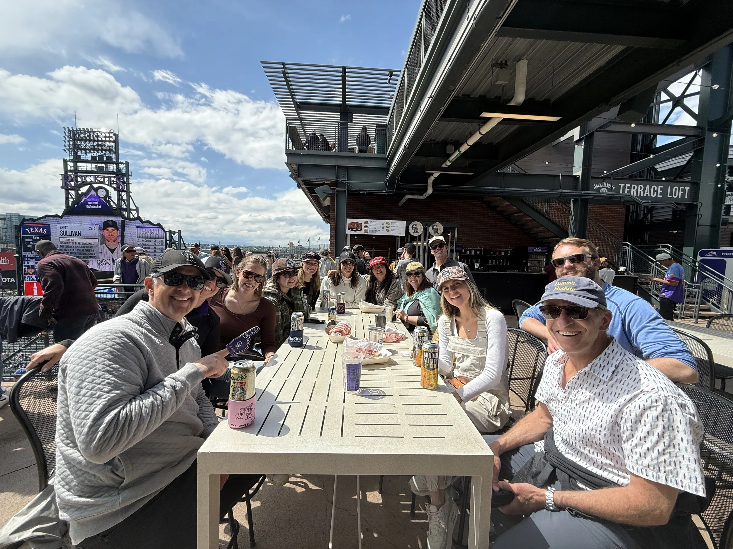 Group of people sitting around a long outdoor table on a rooftop terrace at a baseball stadium, with a large scoreboard and spectators in the background on a partly cloudy day.