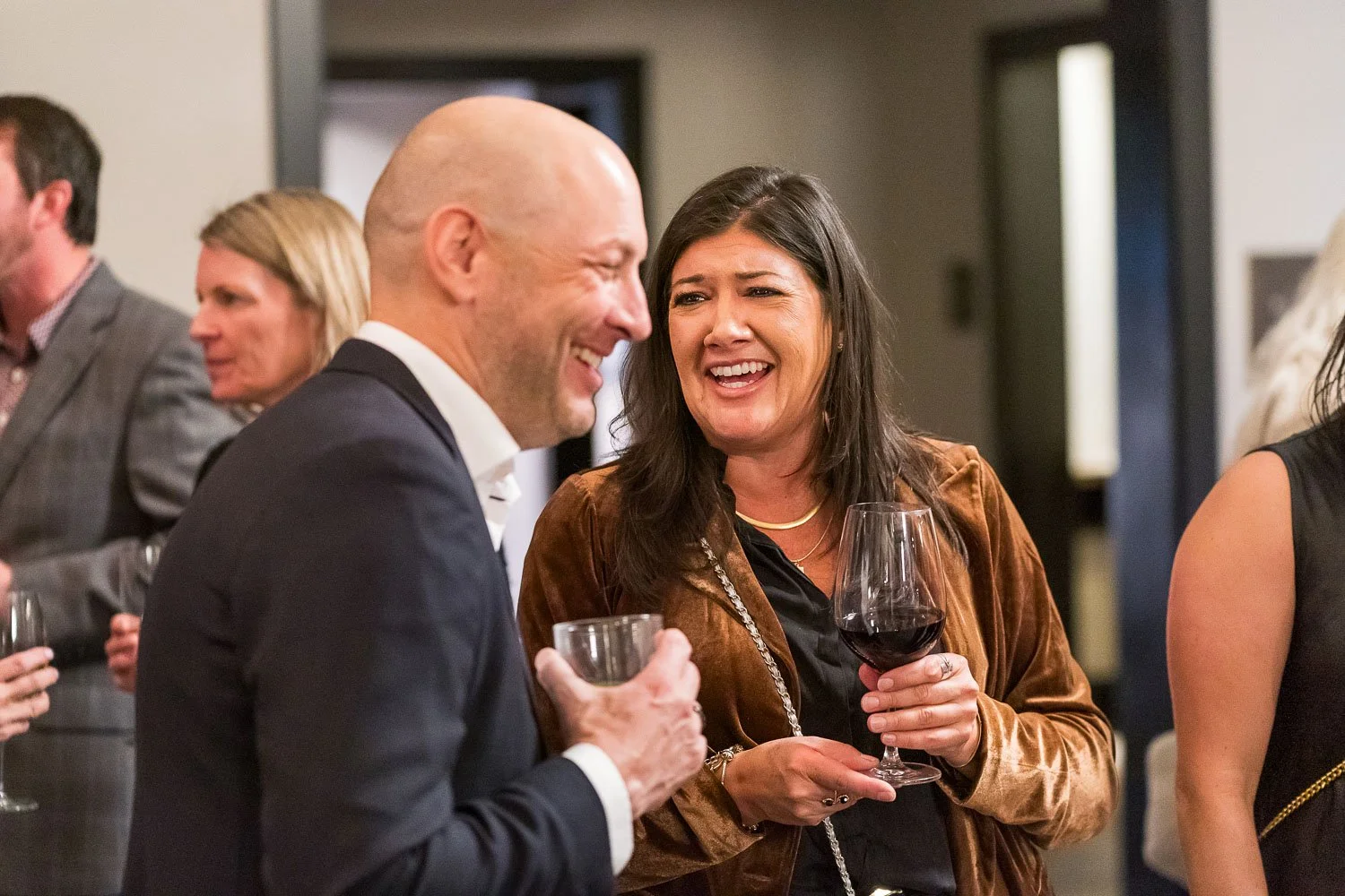 People at a social event are talking and laughing, holding drinks in a warmly lit indoor setting.