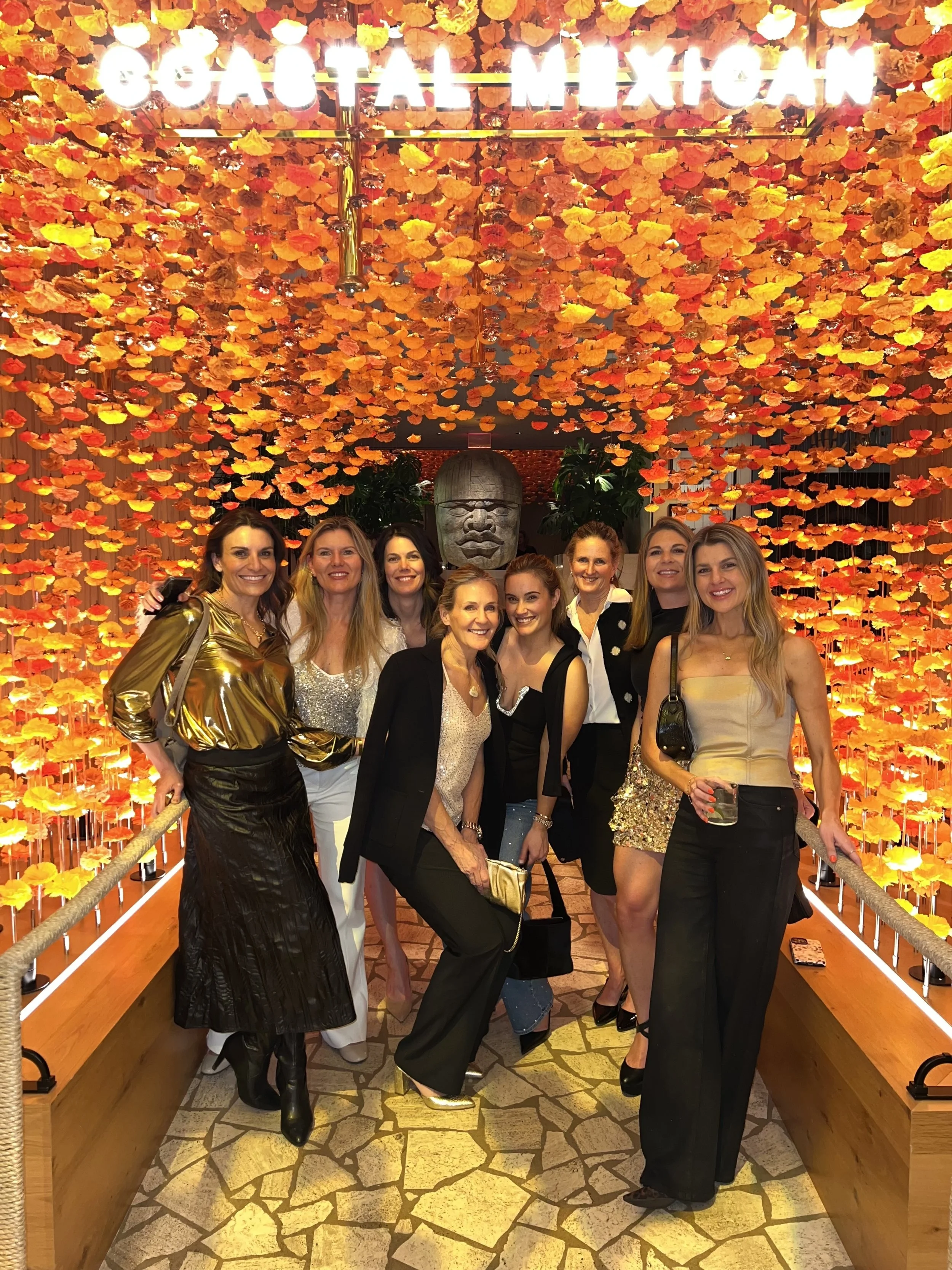 A group of women inside a Mexican-themed restaurant with a glowing sign that reads 'GEOAL MEXICAN' at the top. The ceiling is decorated with vibrant orange and yellow paper flowers, and there is a carved stone face sculpture in the background. The wo