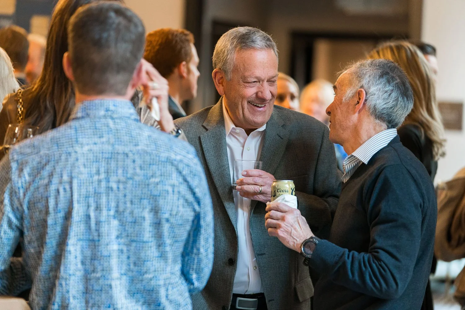 Group of people socializing at a indoor event, including two men in conversation with drinks, smiling and engaging with others in the background.