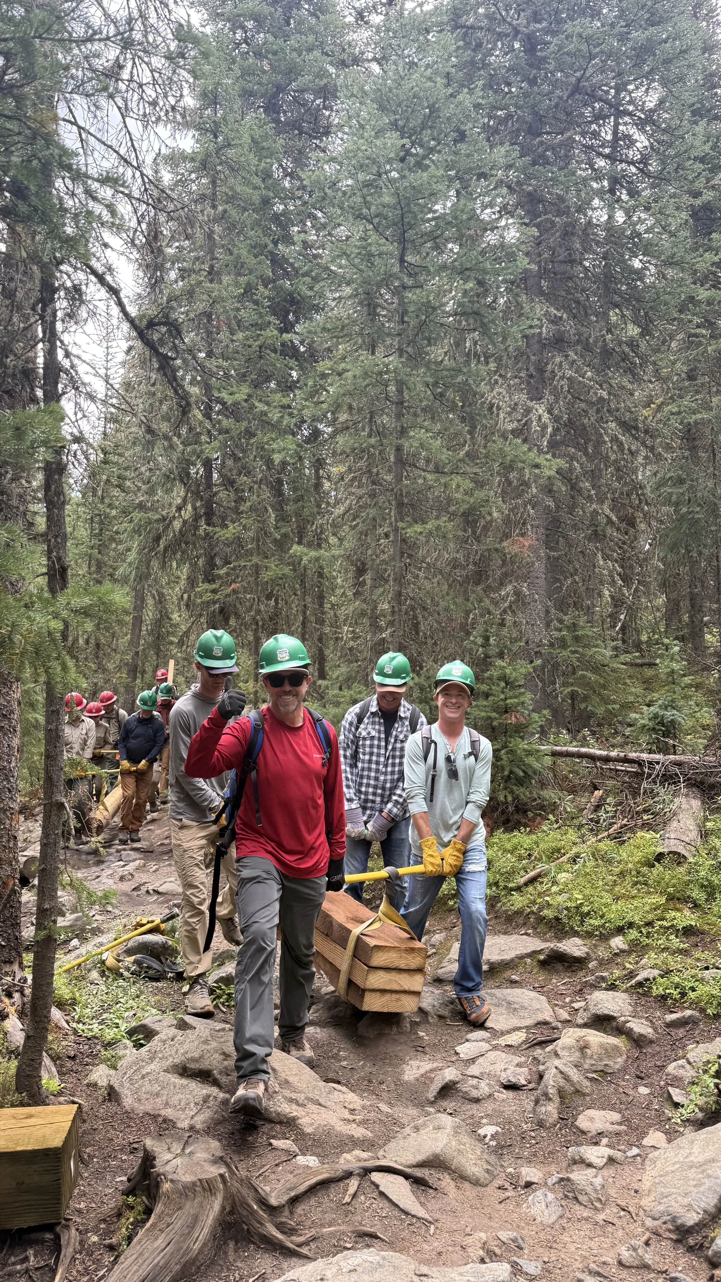 A group of people wearing hard hats and outdoor clothing hiking through a forest. The person in front is smiling and holding a piece of wood with gloves, while others follow along a rocky trail surrounded by tall trees.